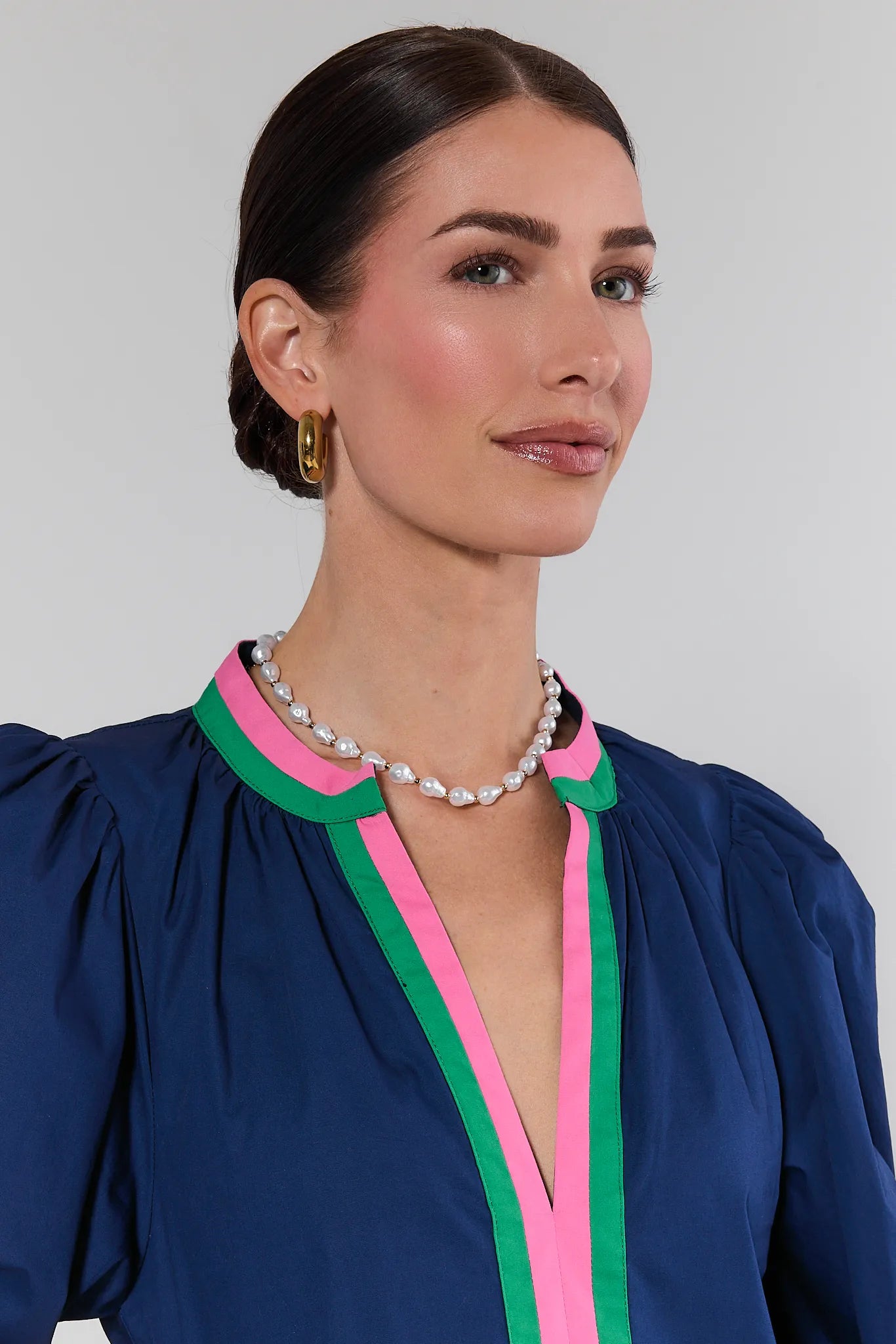 Woman wearing a blue dress with pink and green trim and a pearl necklace against a plain background