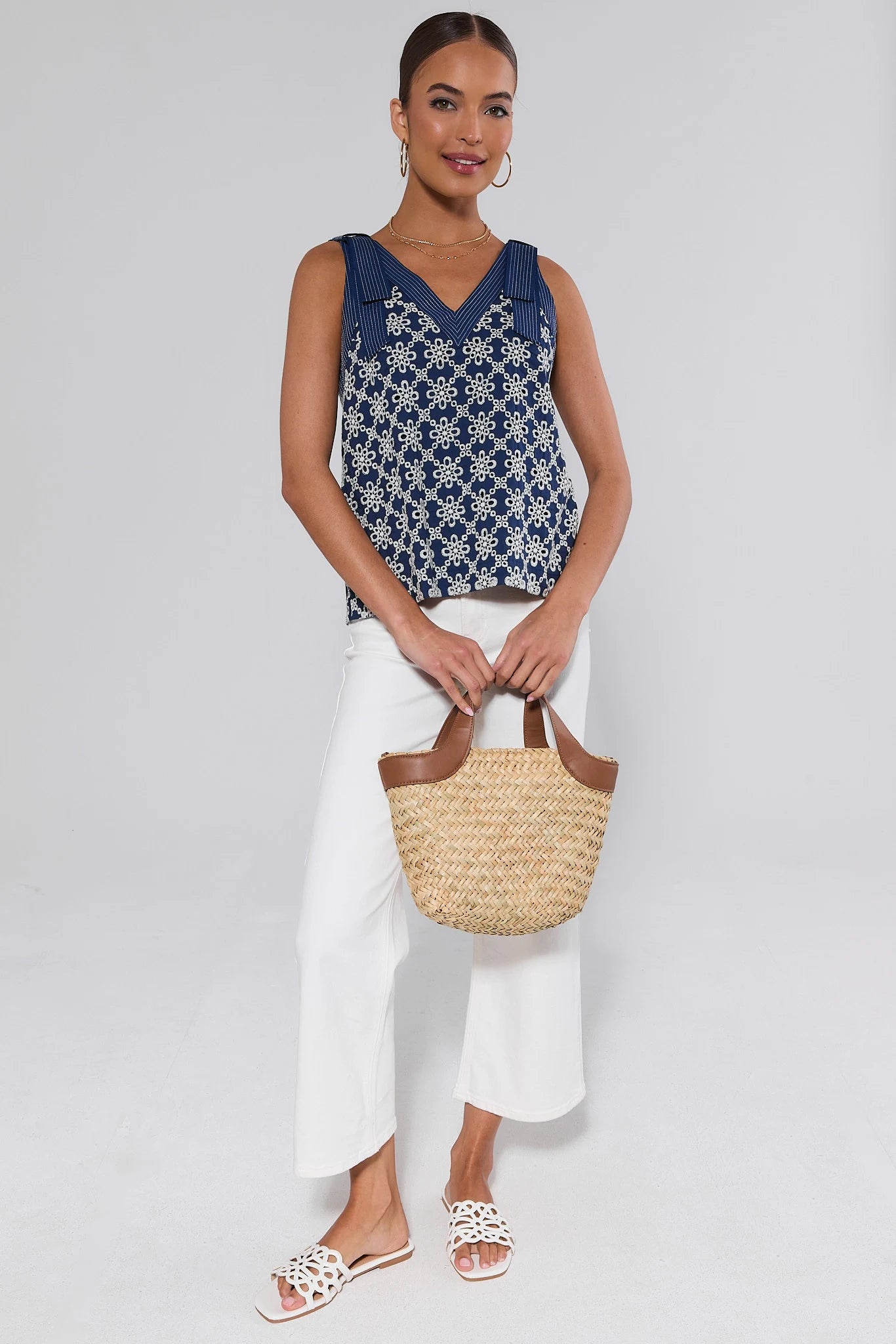 Woman wearing a blue floral top and white pants, holding a straw bag on a light gray background