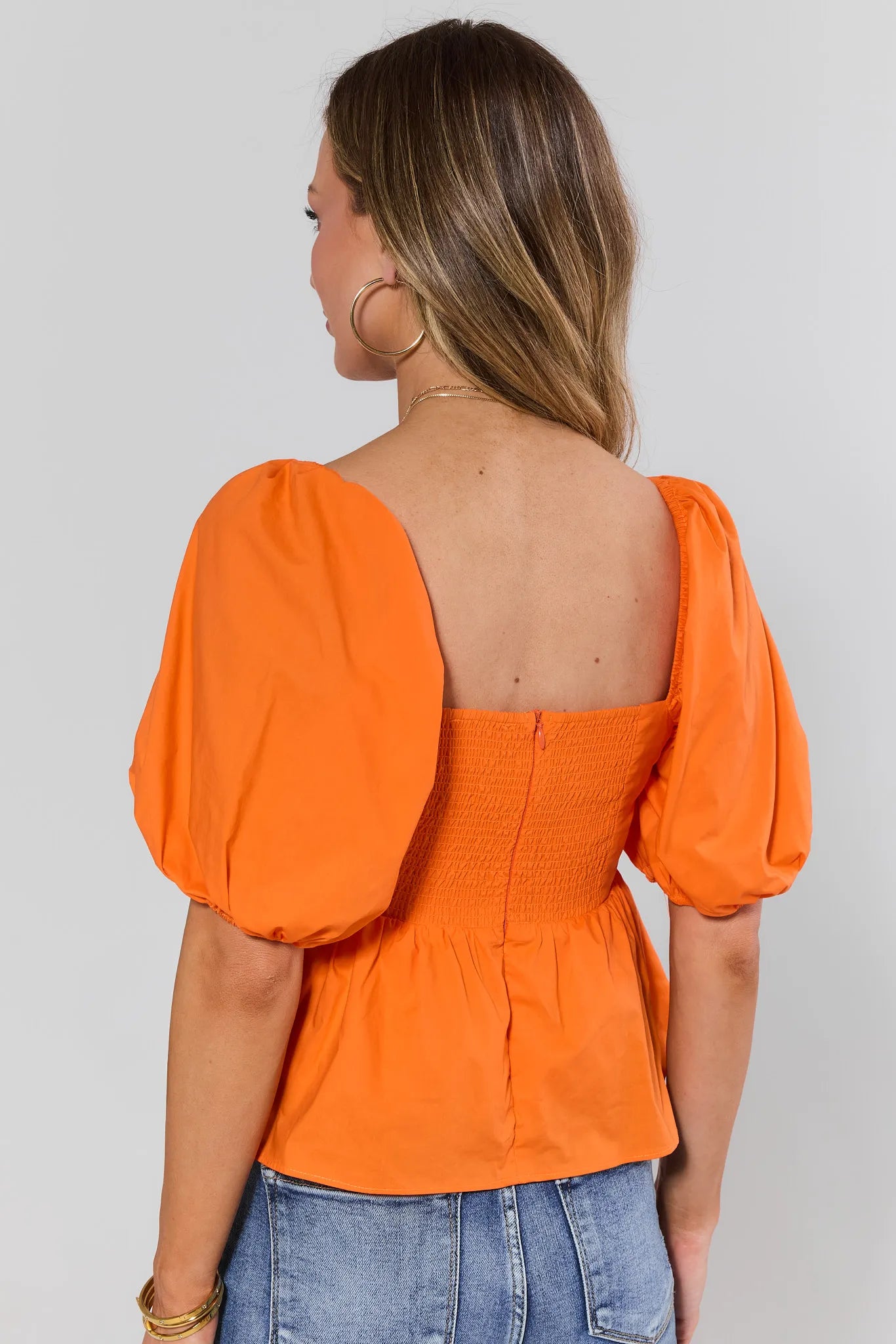 Woman standing wearing an orange top with puffed sleeves on a plain background