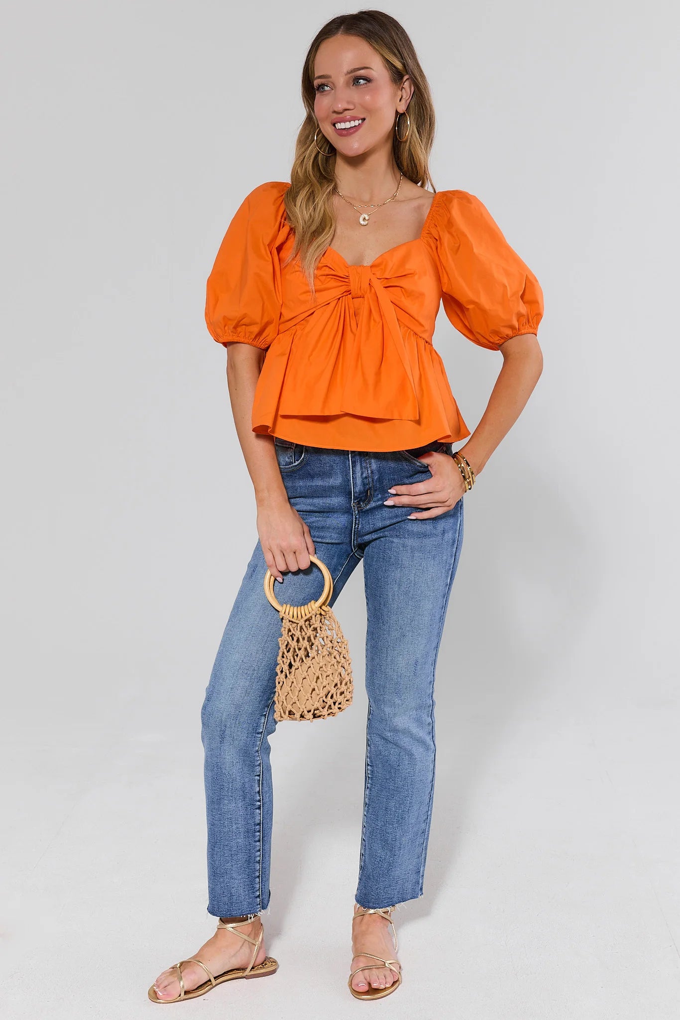 Woman wearing an orange top and blue jeans, holding a woven handbag on a plain background