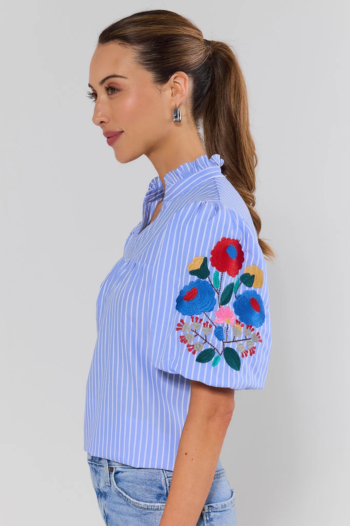 side shot Woman wearing a blue striped shirt with floral embroidery on a plain background