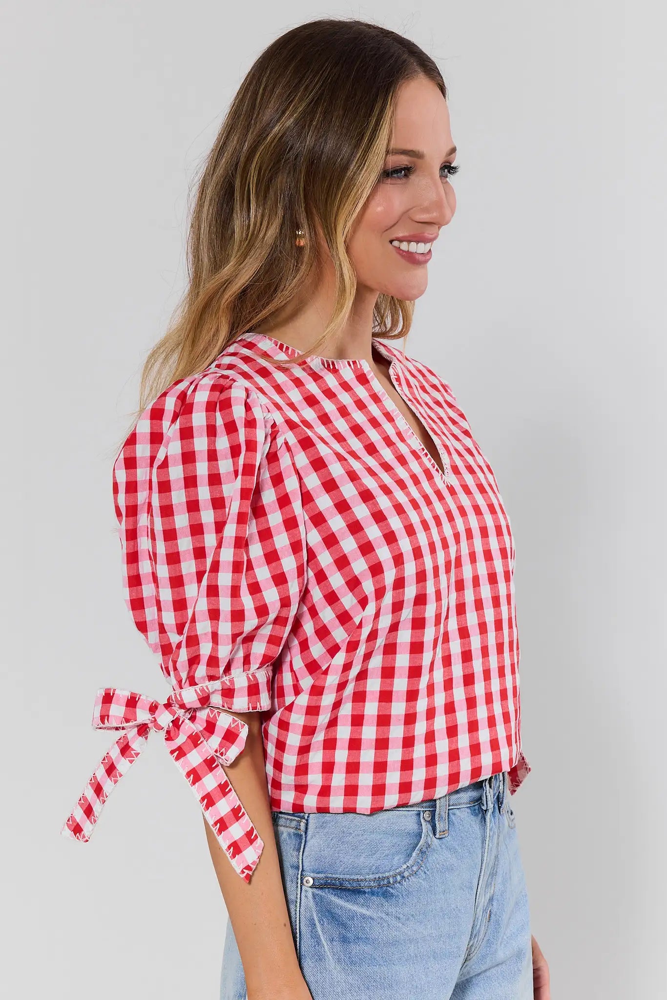 Woman wearing a red and white checkered blouse with tied sleeves against a plain background side shot 