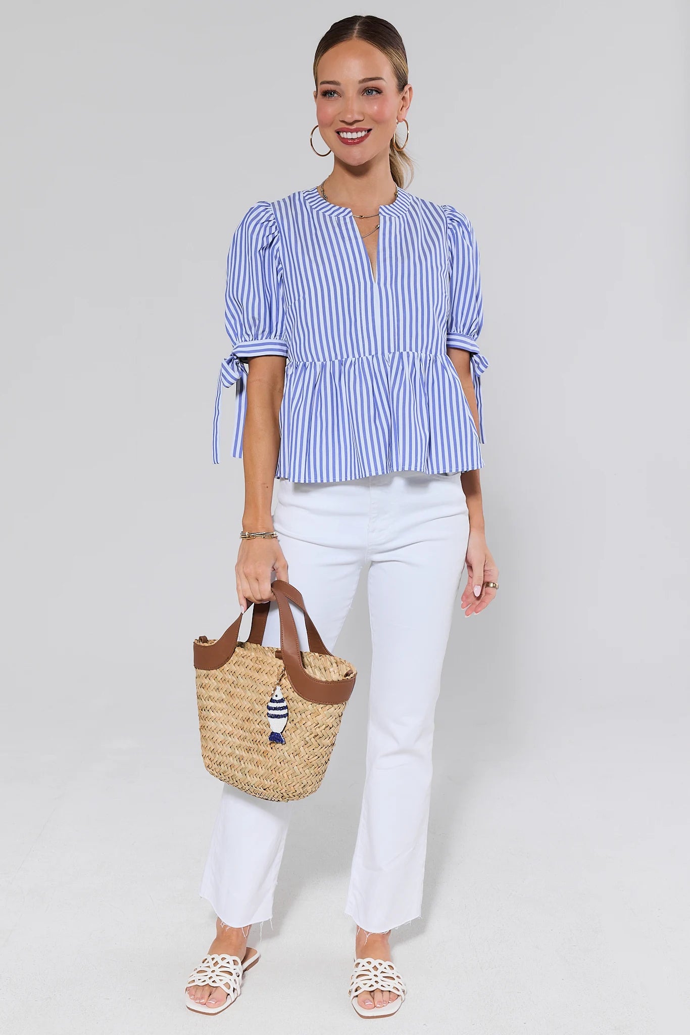 Woman wearing a blue and white striped blouse and white pants, holding a straw bag with a decorative element.