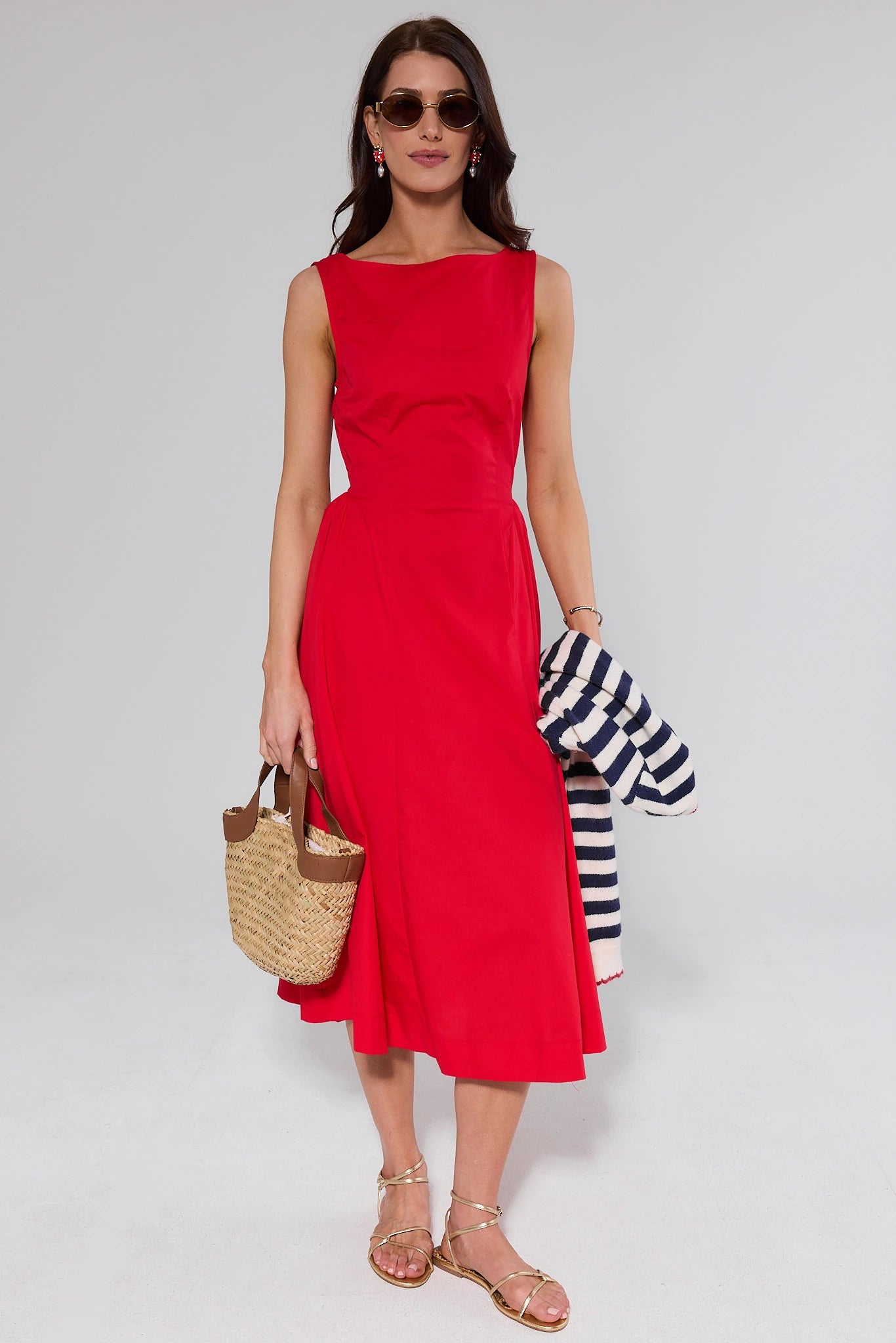 Woman wearing a red dress holding a straw bag and striped shirt on a plain background