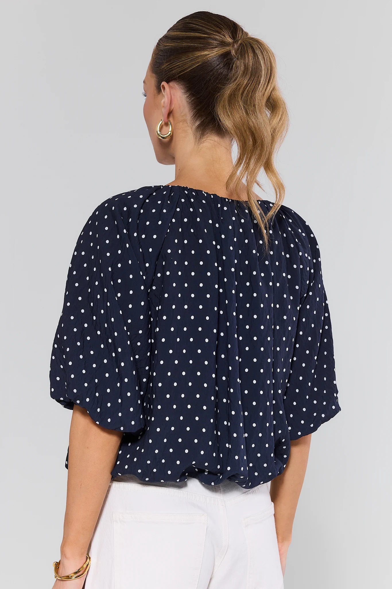 Woman standing wearing a navy blue polka dot blouse against a light gray background