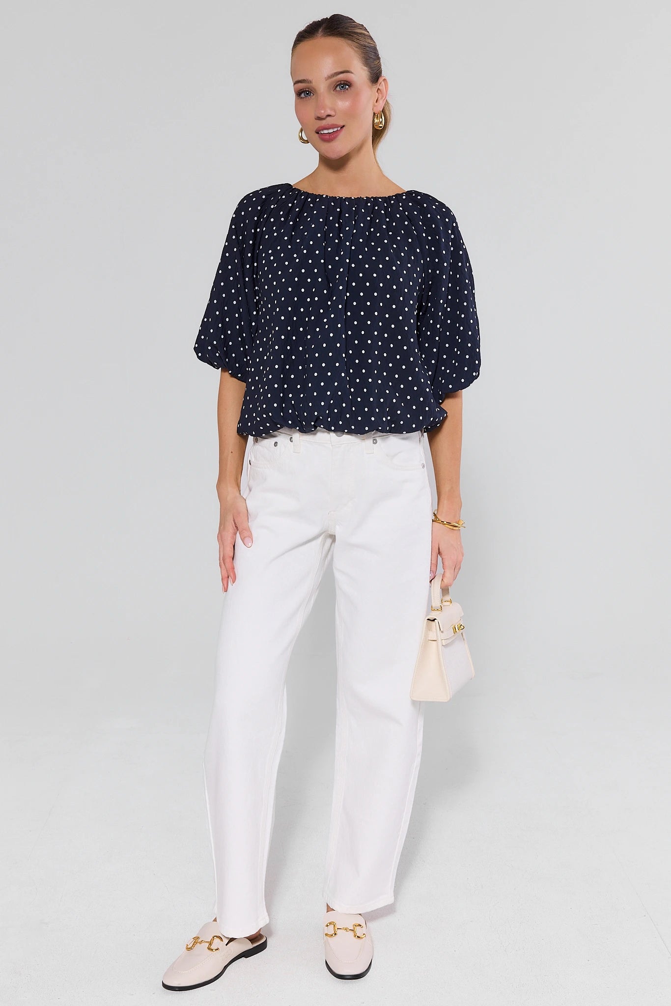 Woman wearing a navy polka dot blouse and white pants against a light gray background