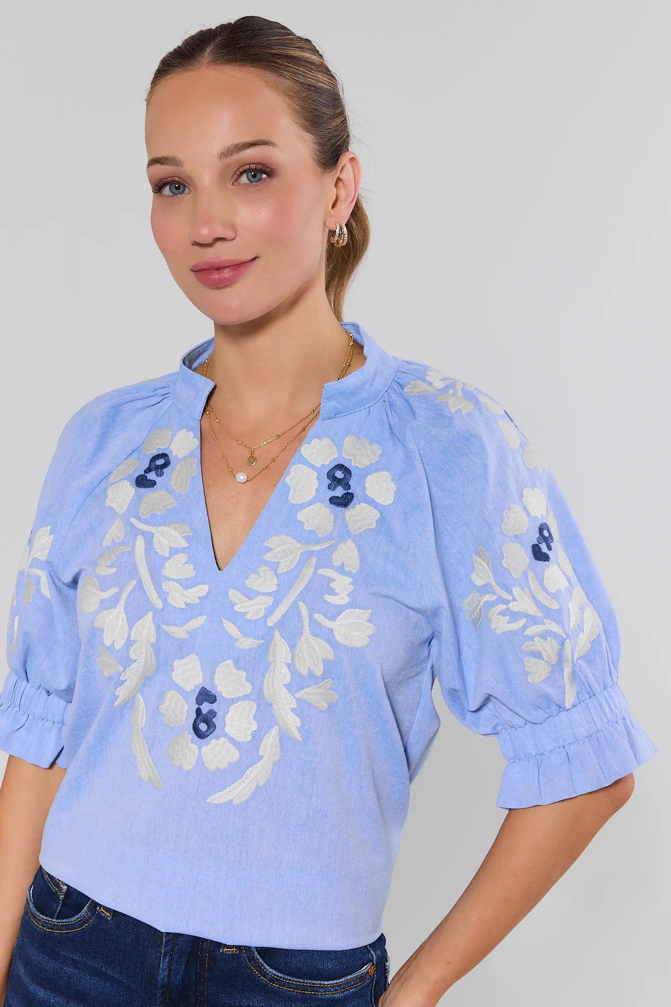 Woman wearing a light blue blouse with white floral embroidery on a plain background