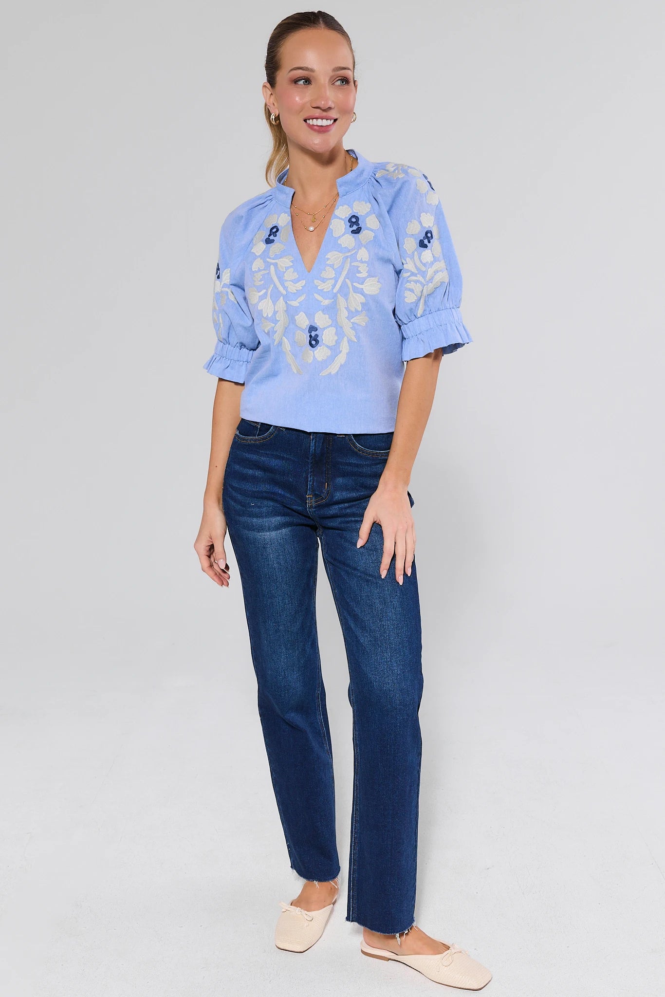Woman wearing a light blue embroidered blouse and dark blue jeans on a gray background