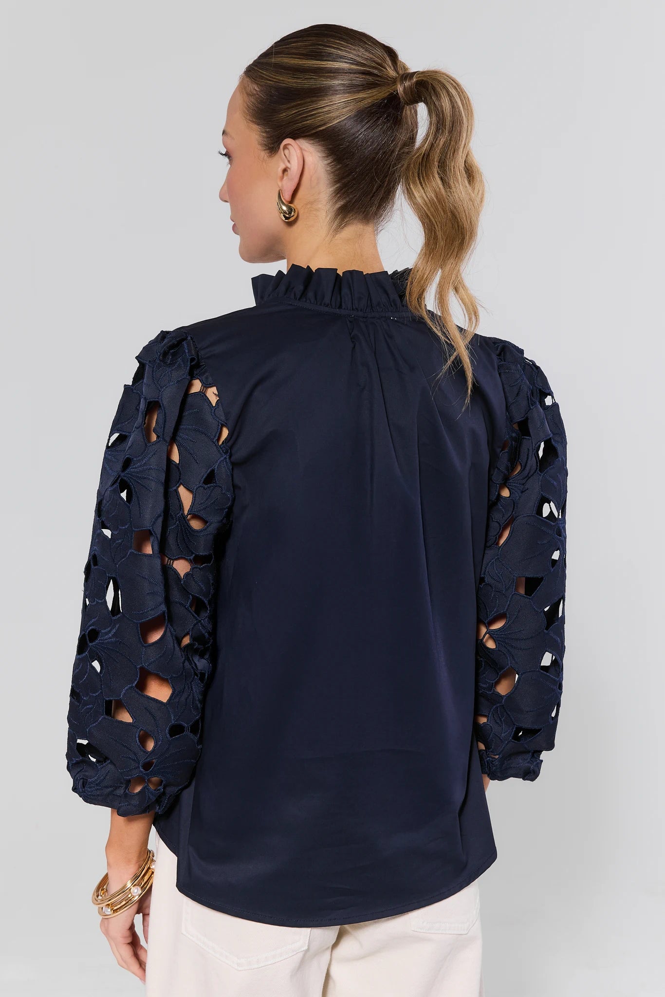 Back view of the Navy Eyelet Sleeve Top by THML, featuring cut-out lace sleeves, a ruffled mock neckline, and a relaxed fit, worn with gold accessories.