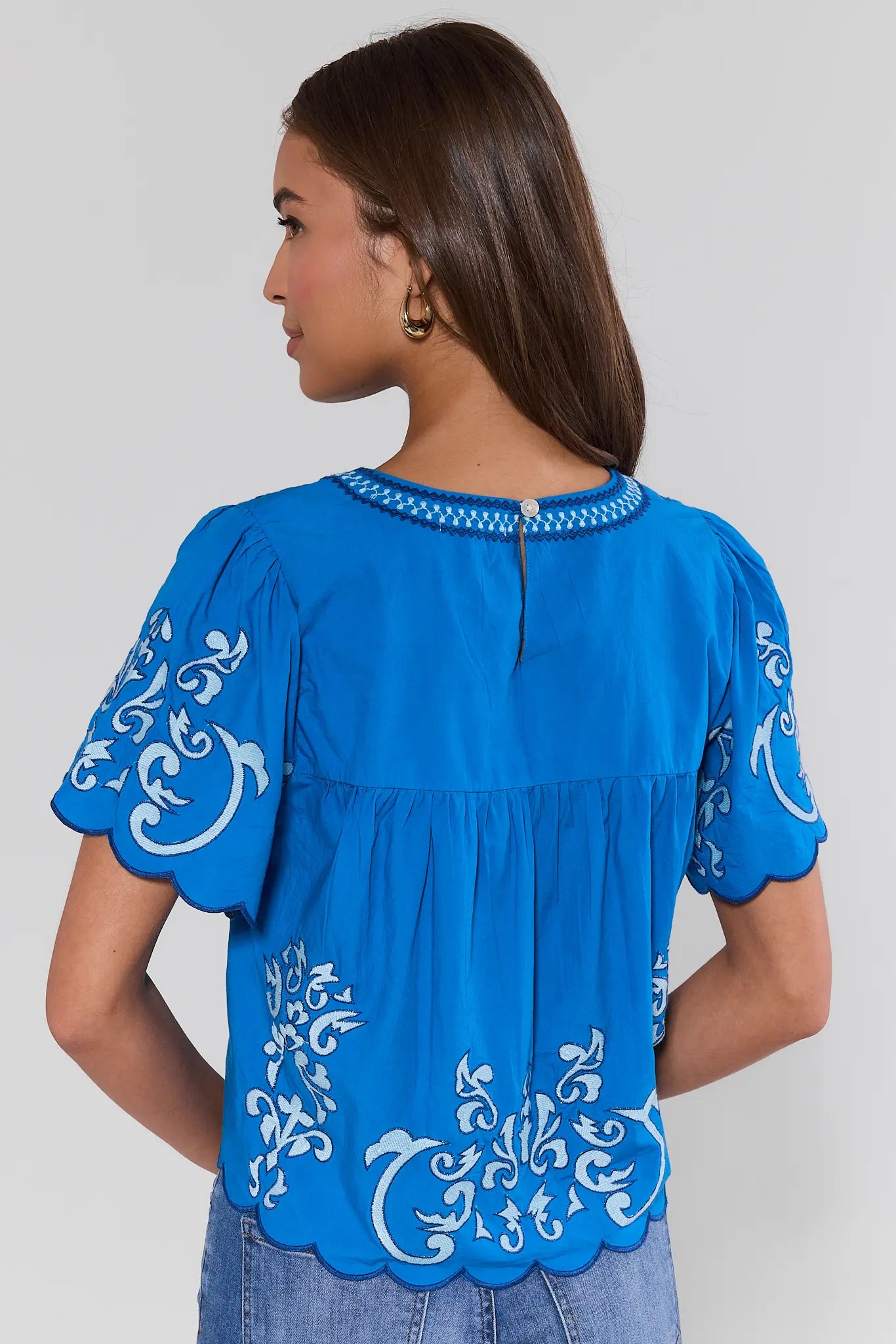 Woman standing wearing a blue blouse with white embroidery on a plain background