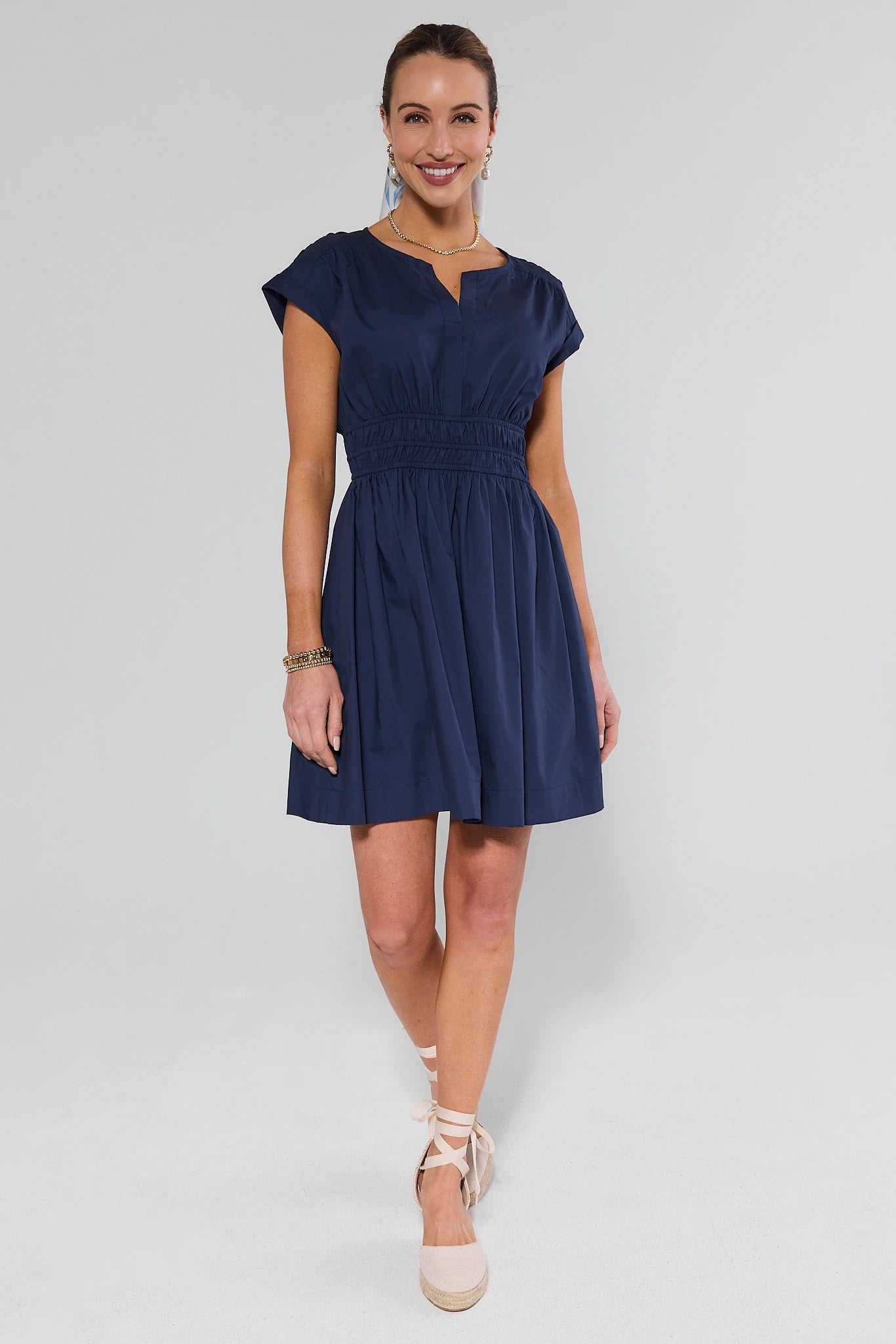 Woman wearing Navy Smocked Waist Short Dress with gathered waist, a-line skirt, notched v-neckline, cap sleeves, and espadrille wedges, accessorized with jewelry.