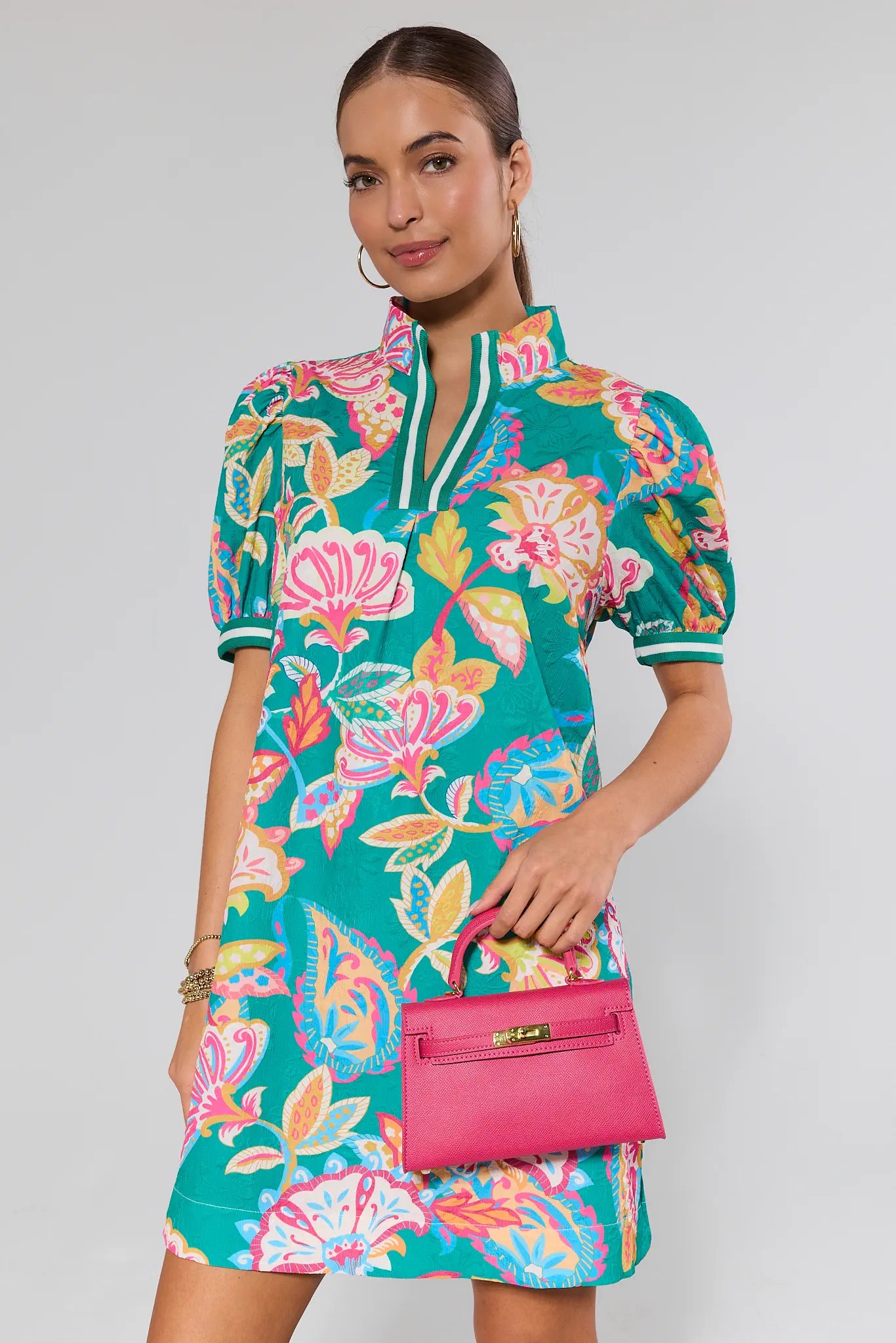 Woman wearing a colorful floral dress holding a pink handbag against a background