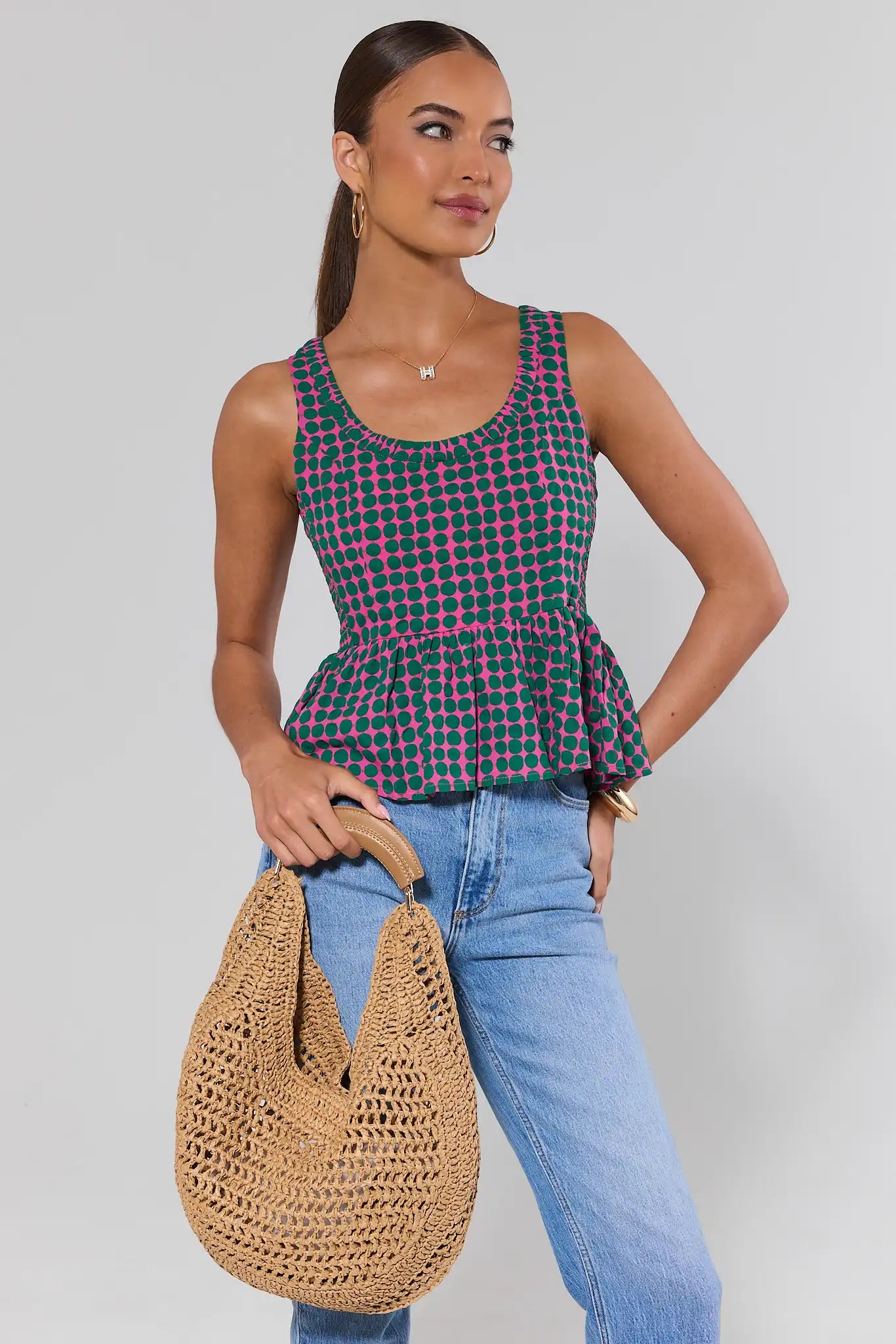 Woman wearing a green and pink checkered top and blue jeans, holding a woven bag against a plain background