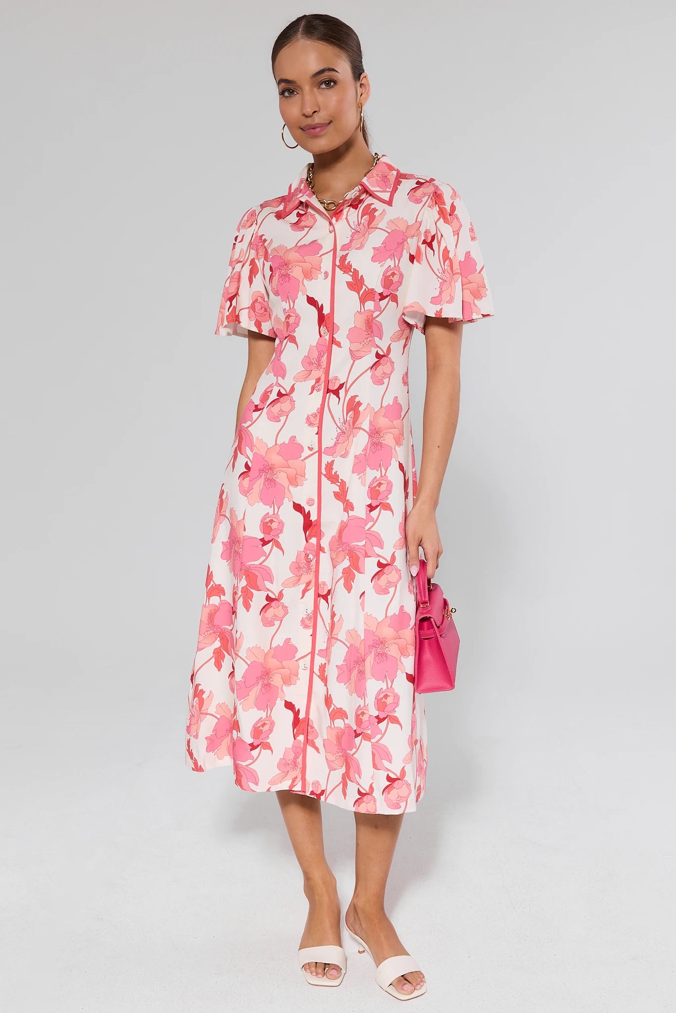 Woman wearing a pink floral dress holding a pink handbag against a gray background