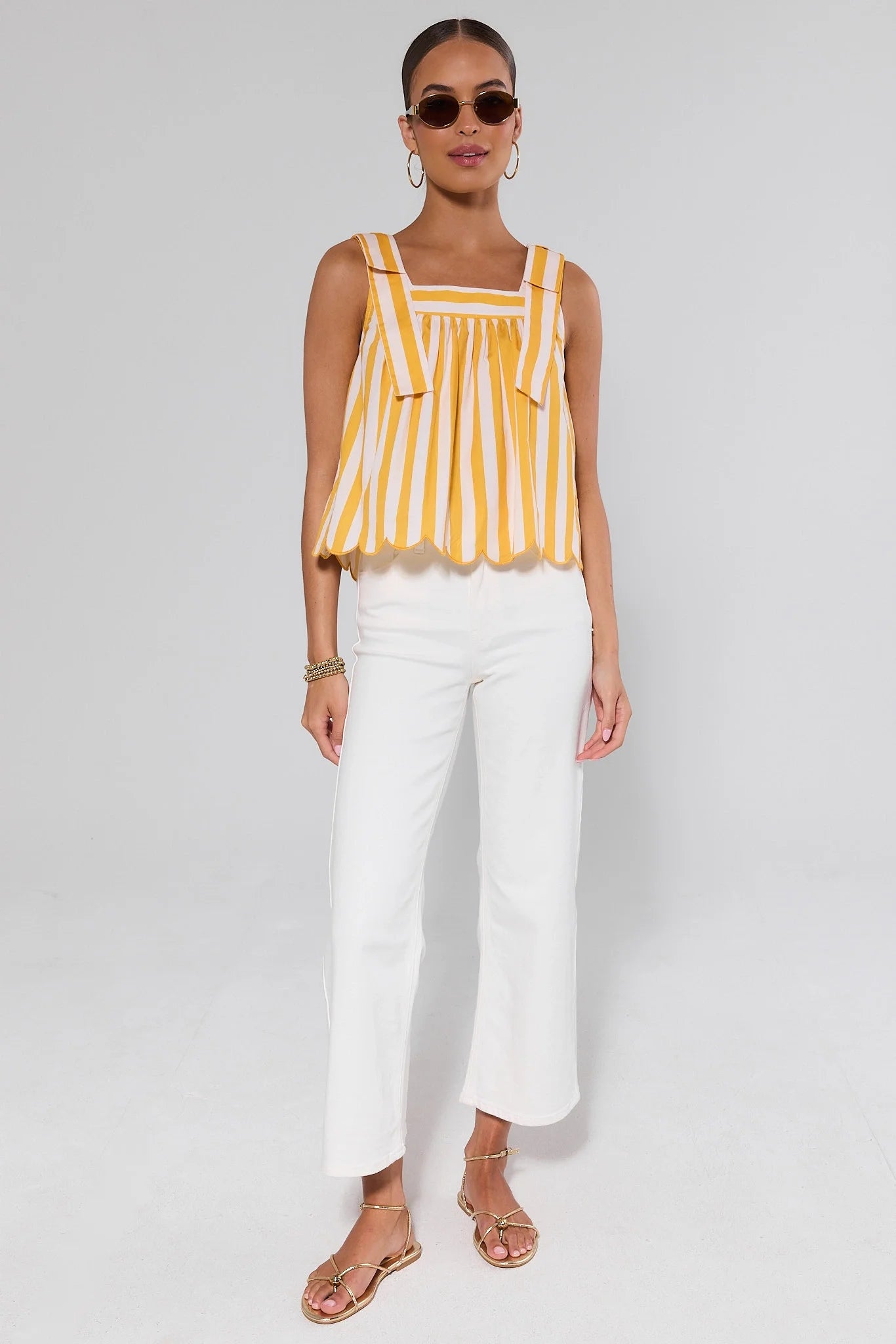 Woman wearing a yellow and white striped sleeveless top with white pants on a plain background