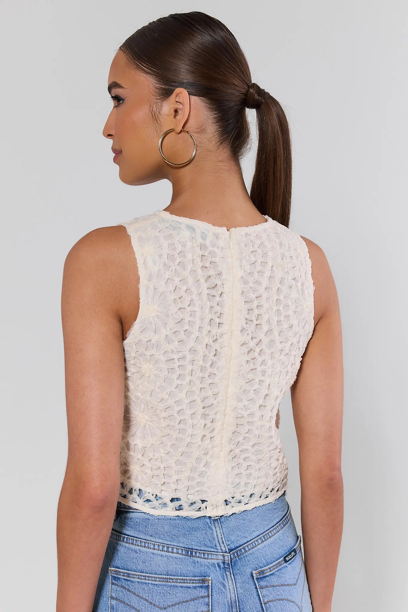 Woman standing wearing a white sleeveless top and blue jeans against a plain background
