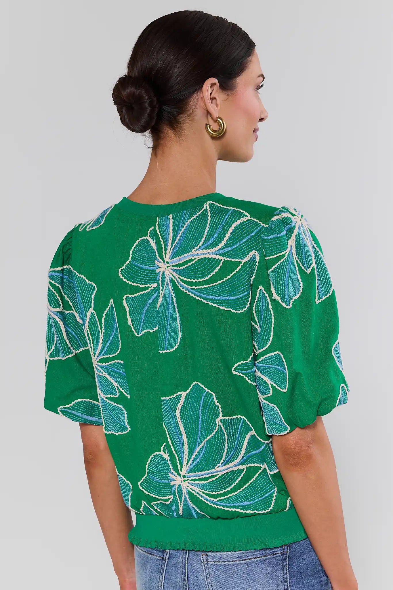 Woman standing wearing a green blouse with floral patterns on a plain background
