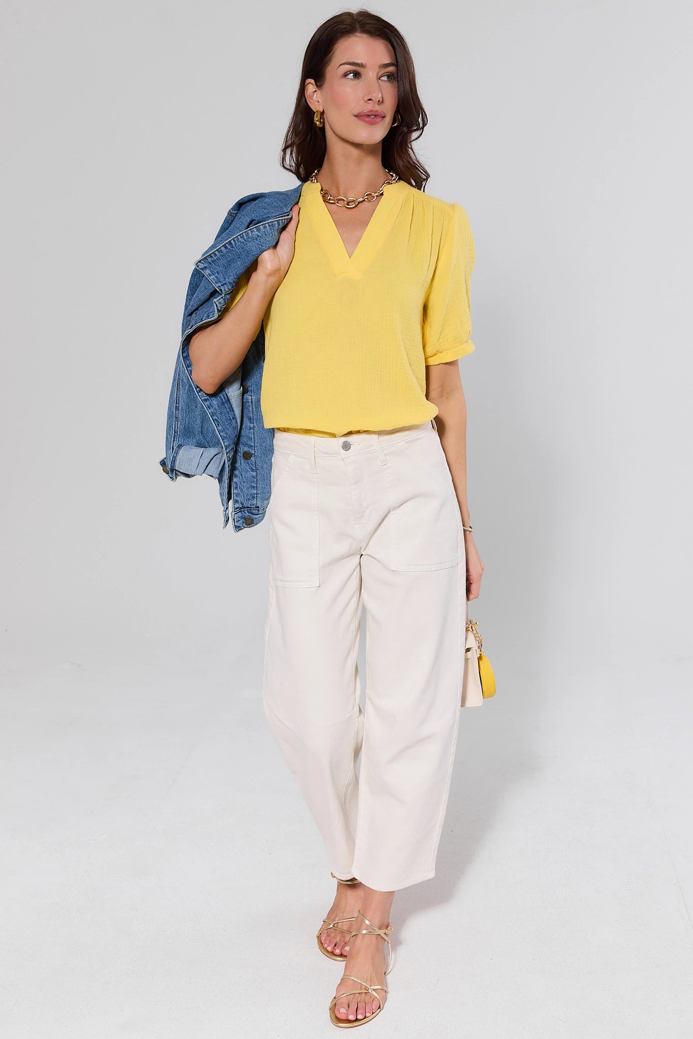 Woman wearing a yellow blouse, white pants, and a denim jacket on a plain background