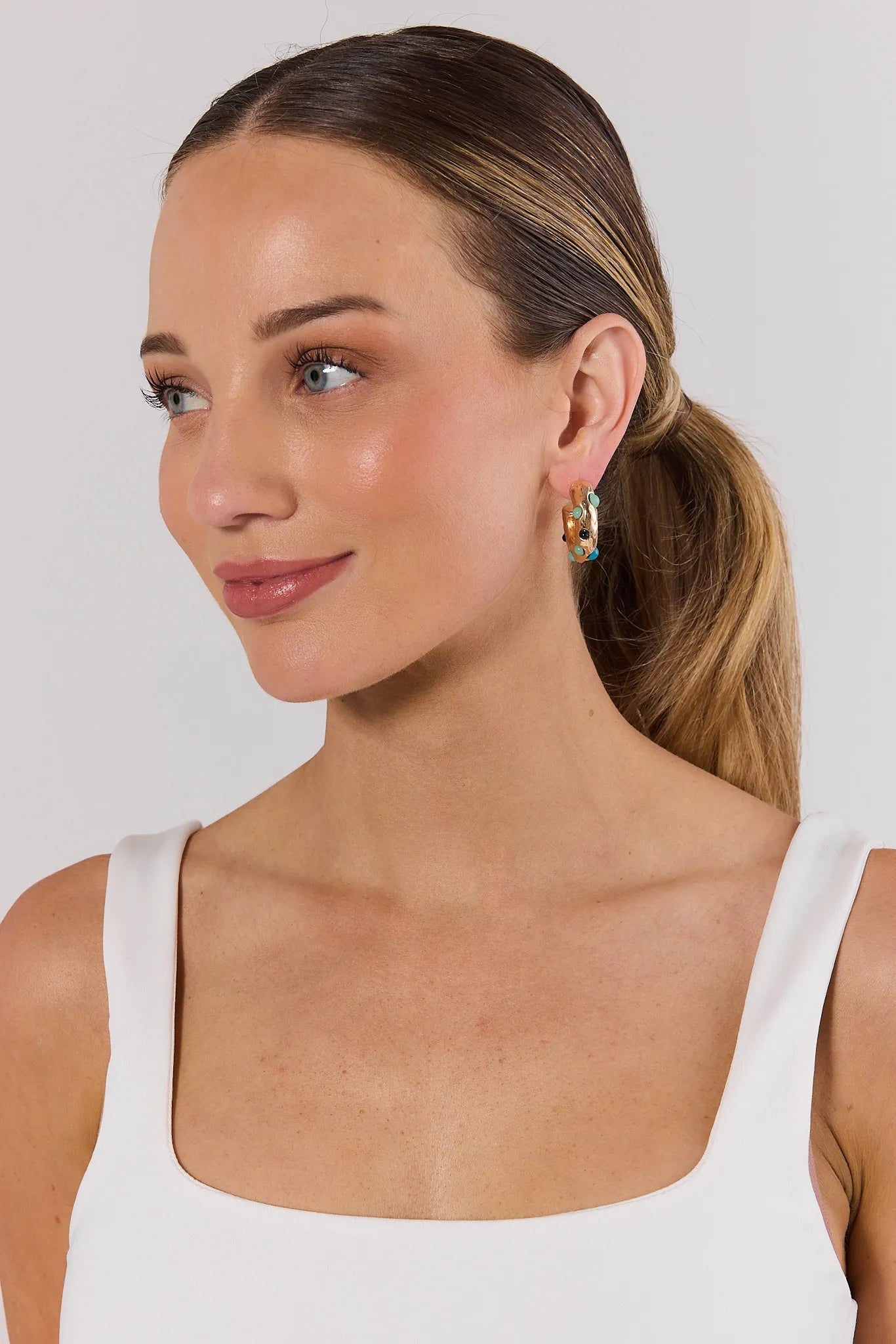Woman wearing a white tank top and multi earrings against a plain background