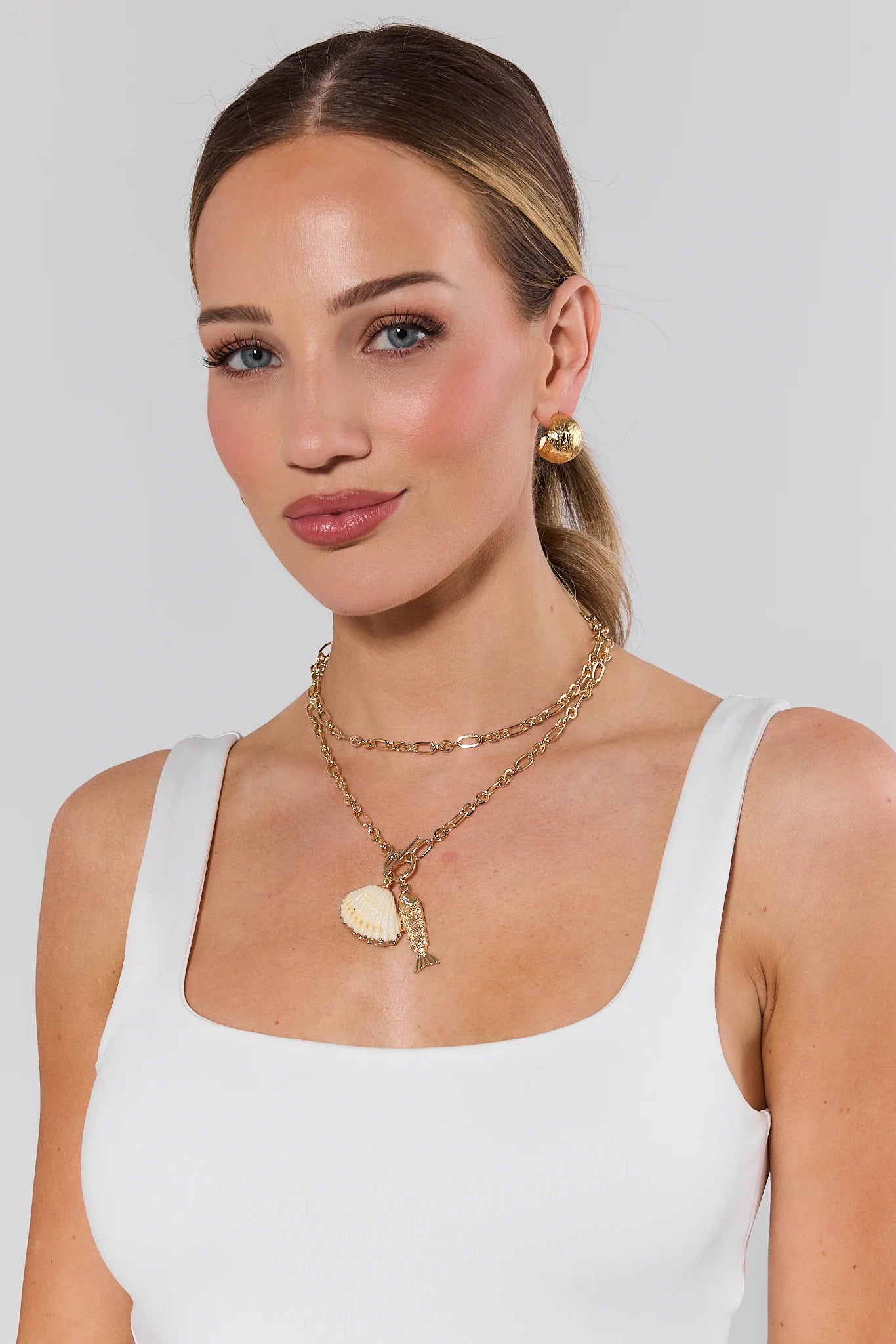 Woman wearing gold shell jewelry including earrings and necklaces on a plain background