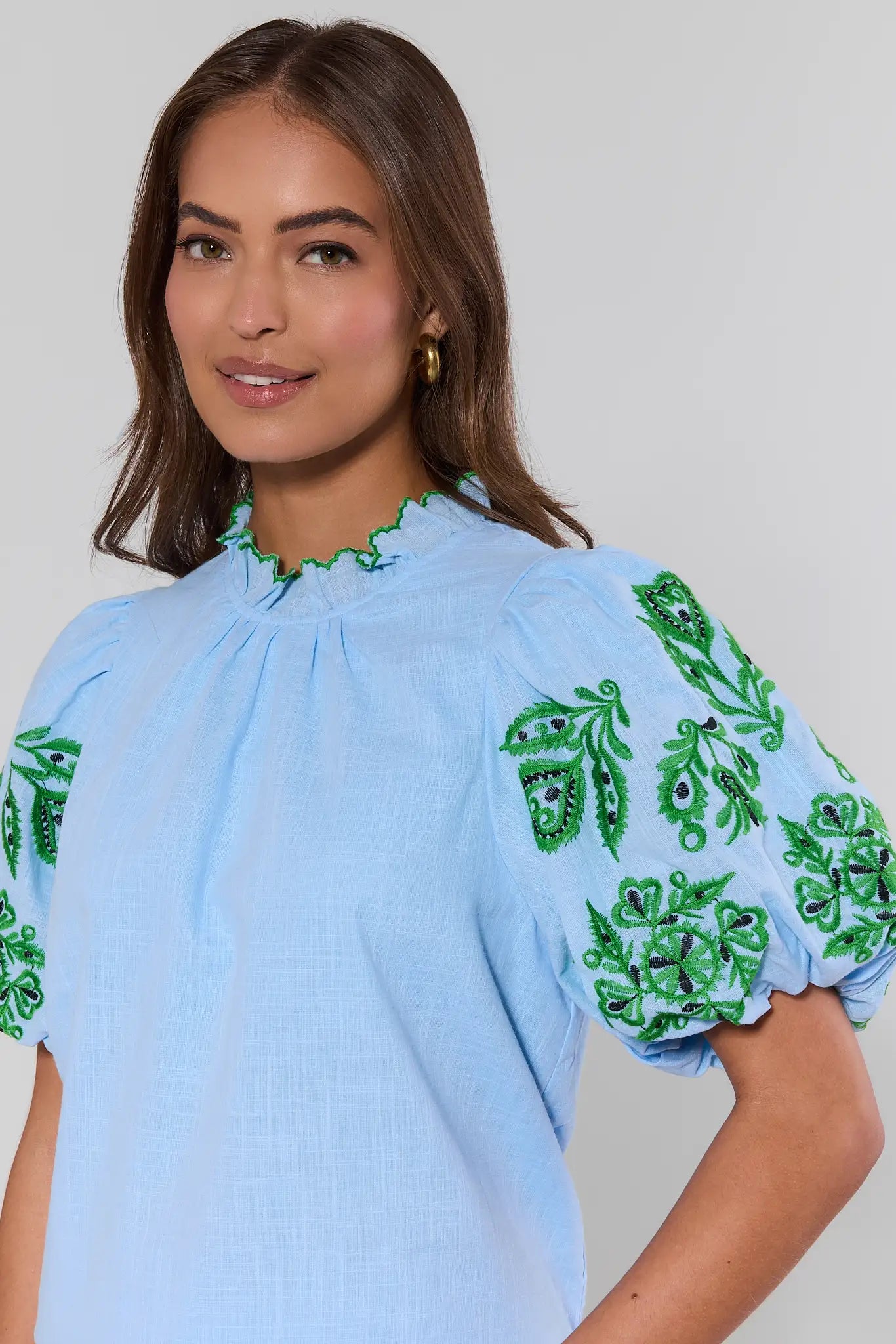 close up Woman wearing a light blue blouse with green embroidery on a plain background