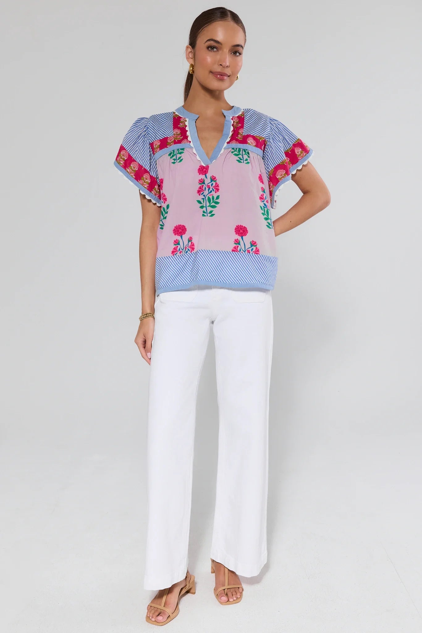 Woman wearing a colorful embroidered top and white pants on a plain background