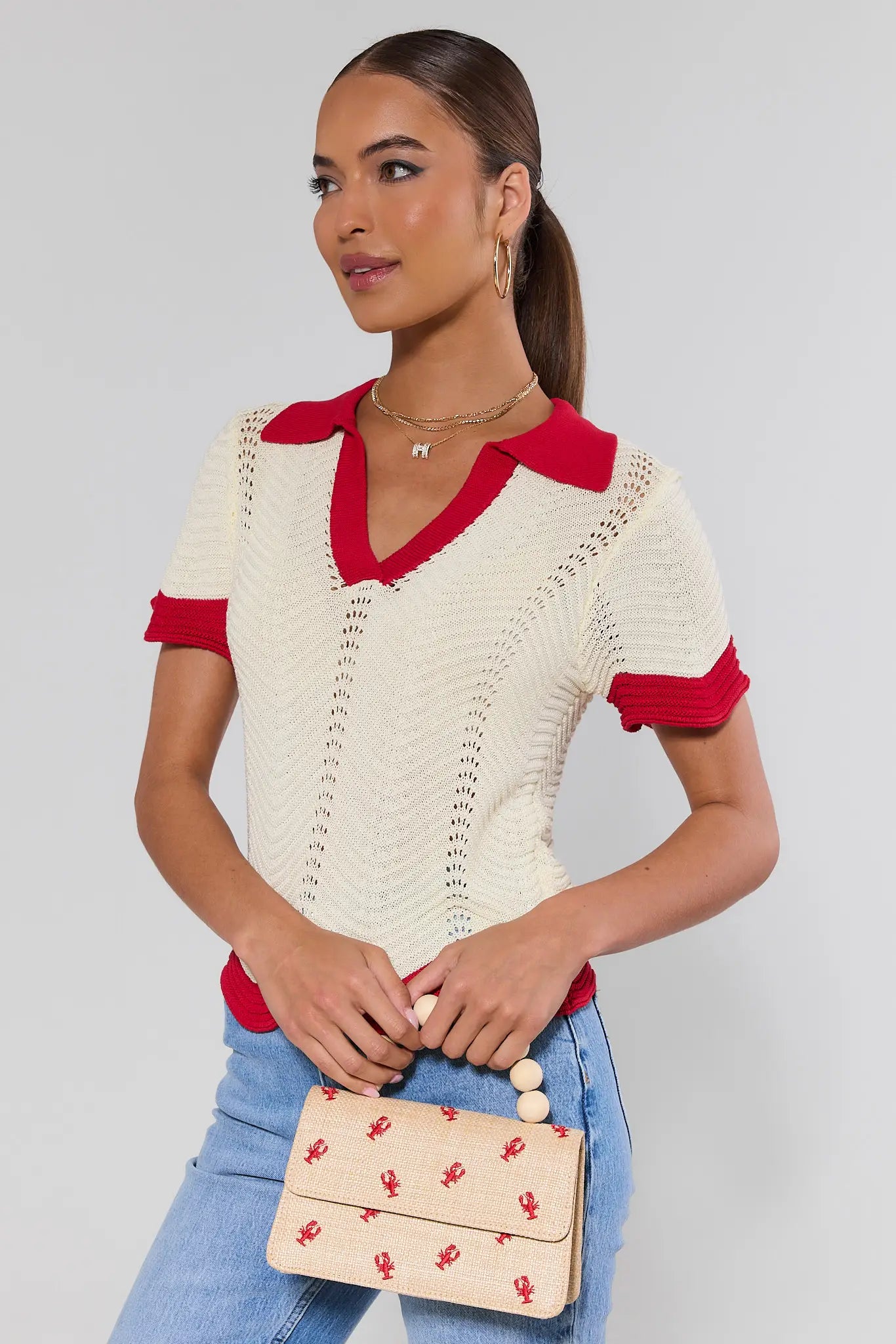 Woman wearing a cream and red top with blue jeans, holding a beige clutch with red patterns.