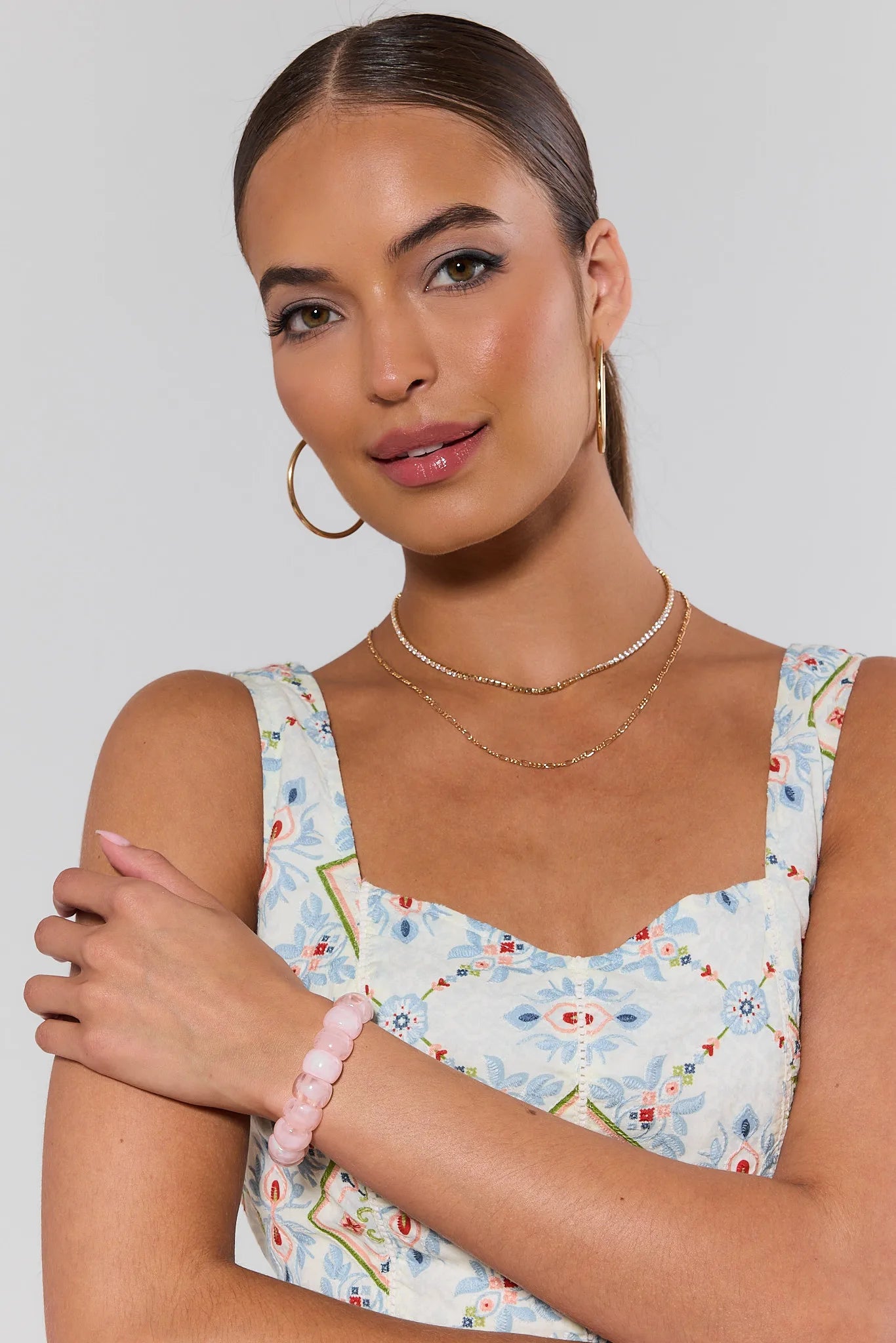 Woman wearing a floral dress with layered necklaces and a pink bracelet on a plain background