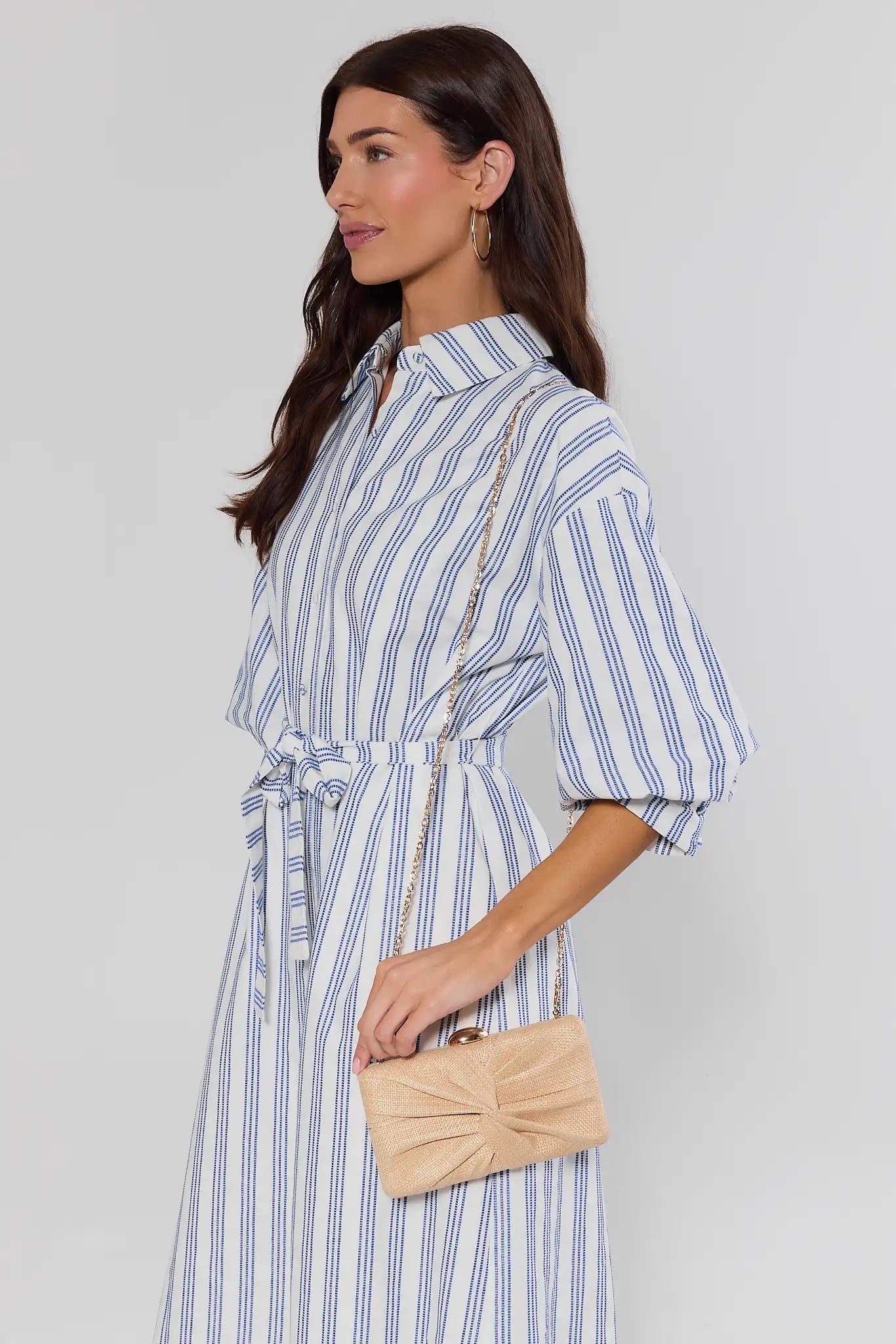 Woman wearing a blue and white striped dress holding a beige clutch against a plain background