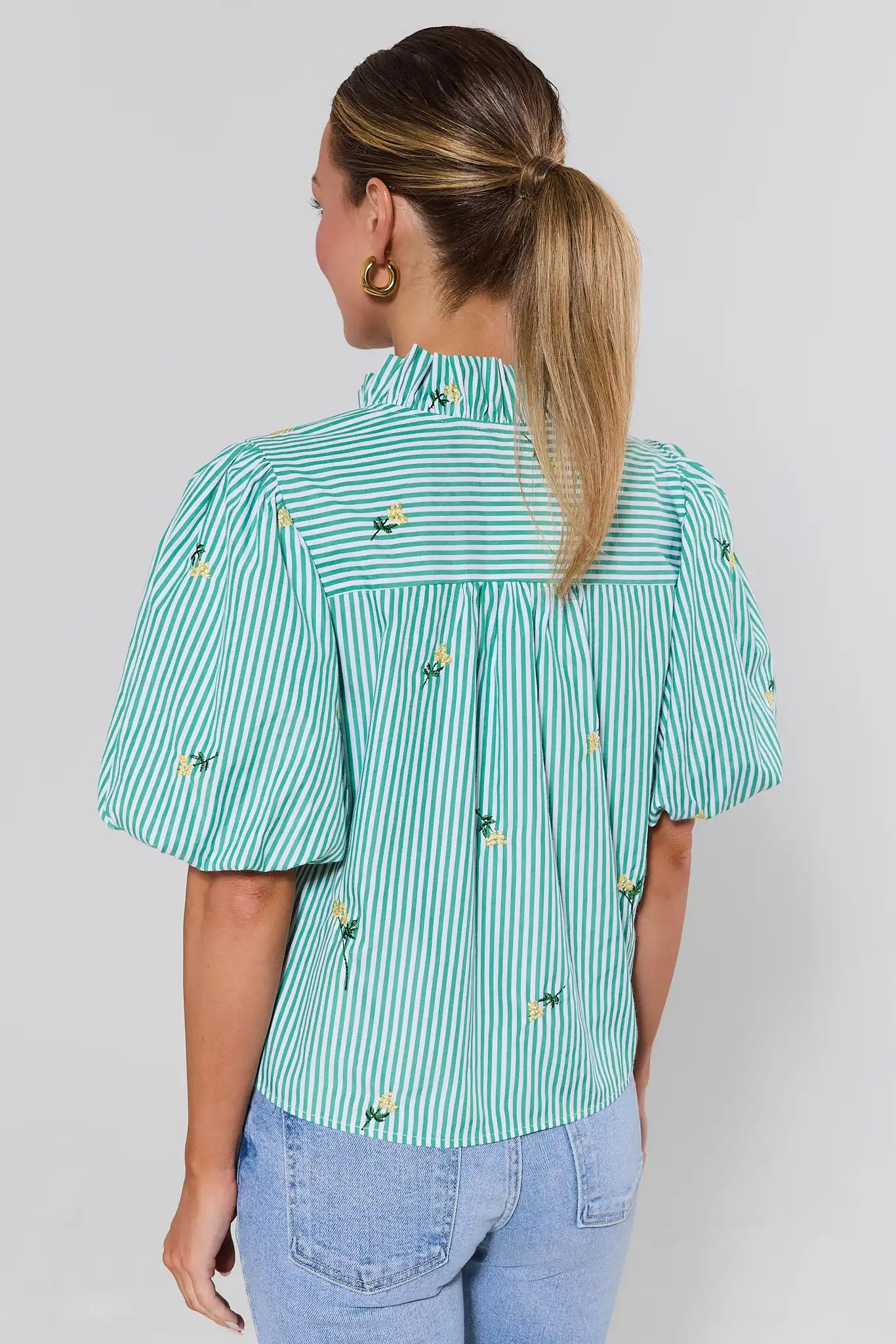 Woman standing wearing a green and white striped shirt with floral patterns on a plain background