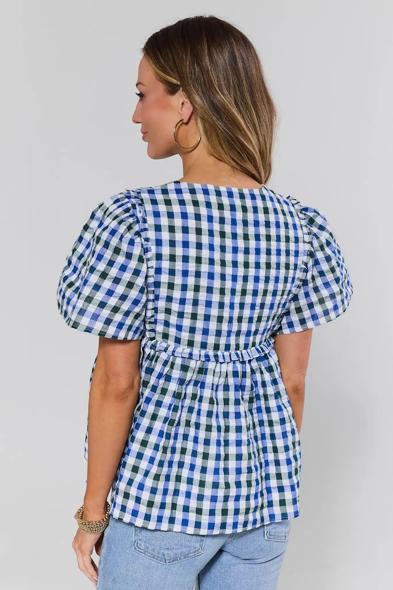 Woman standing wearing a blue and white checkered blouse with puff sleeves on a plain background
