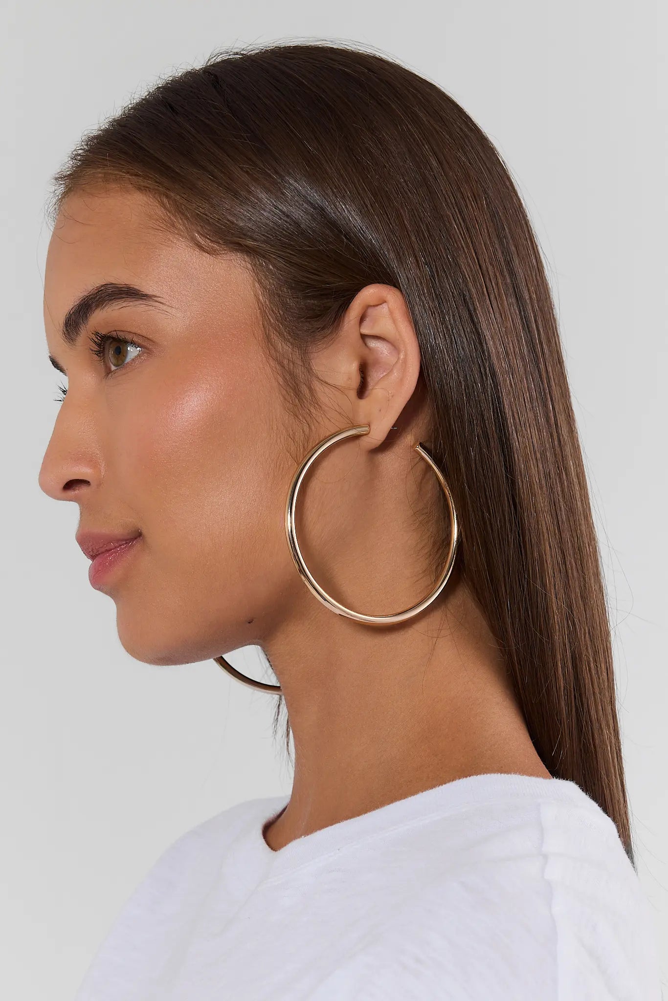 A close-up side profile of a woman with long dark brown hair wearing large, thin gold hoop earrings and a white top, set against a plain light gray background.