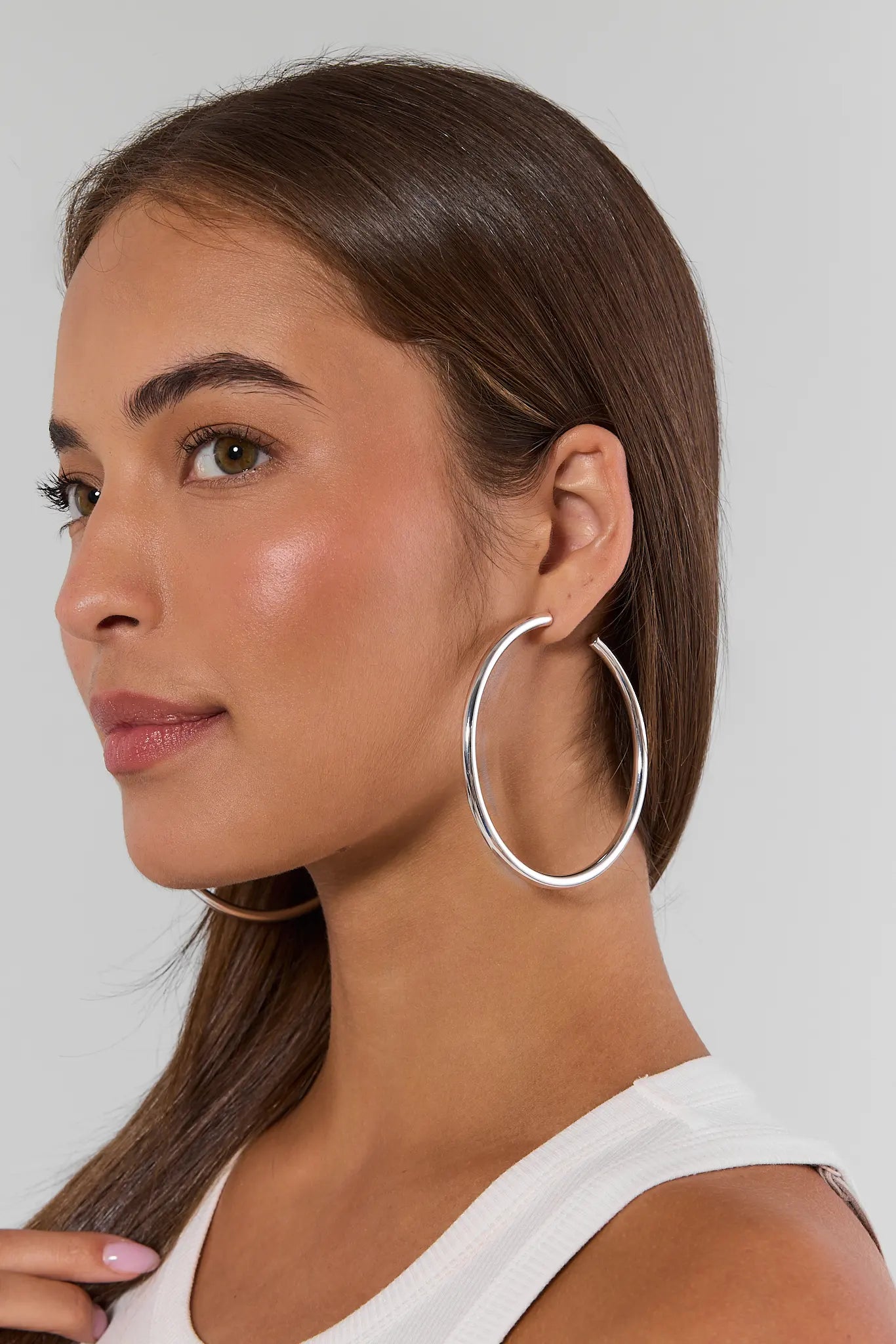 A close-up side profile of a woman wearing a pair of large, polished silver hoop earrings against a light gray background.