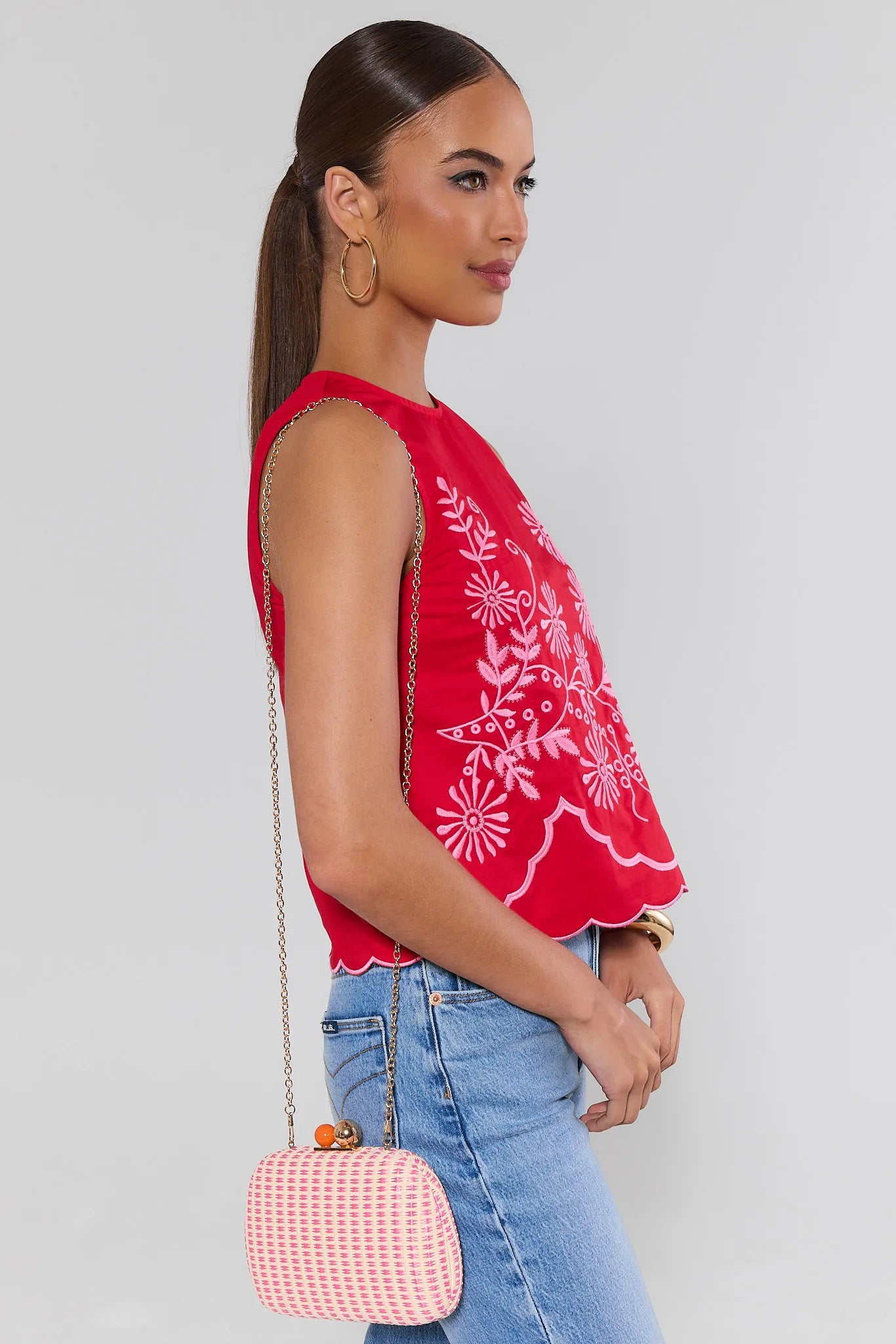 Woman wearing a red embroidered top and blue jeans, holding a pink handbag against a white background
