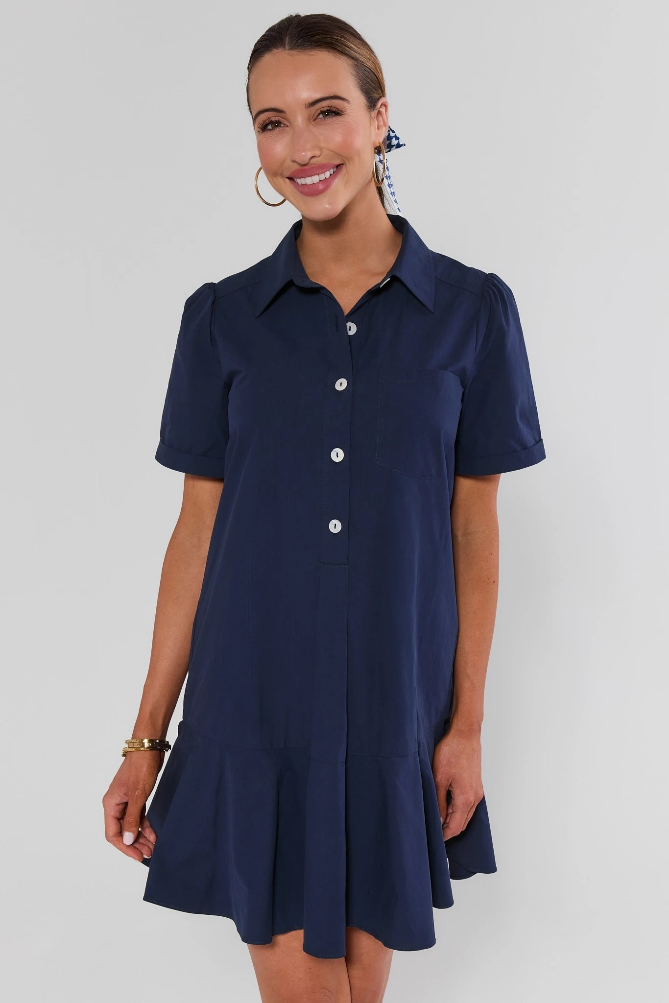 A woman modeling a short-sleeve navy blue collared shirt dress with white buttons, set against a plain light gray studio background.