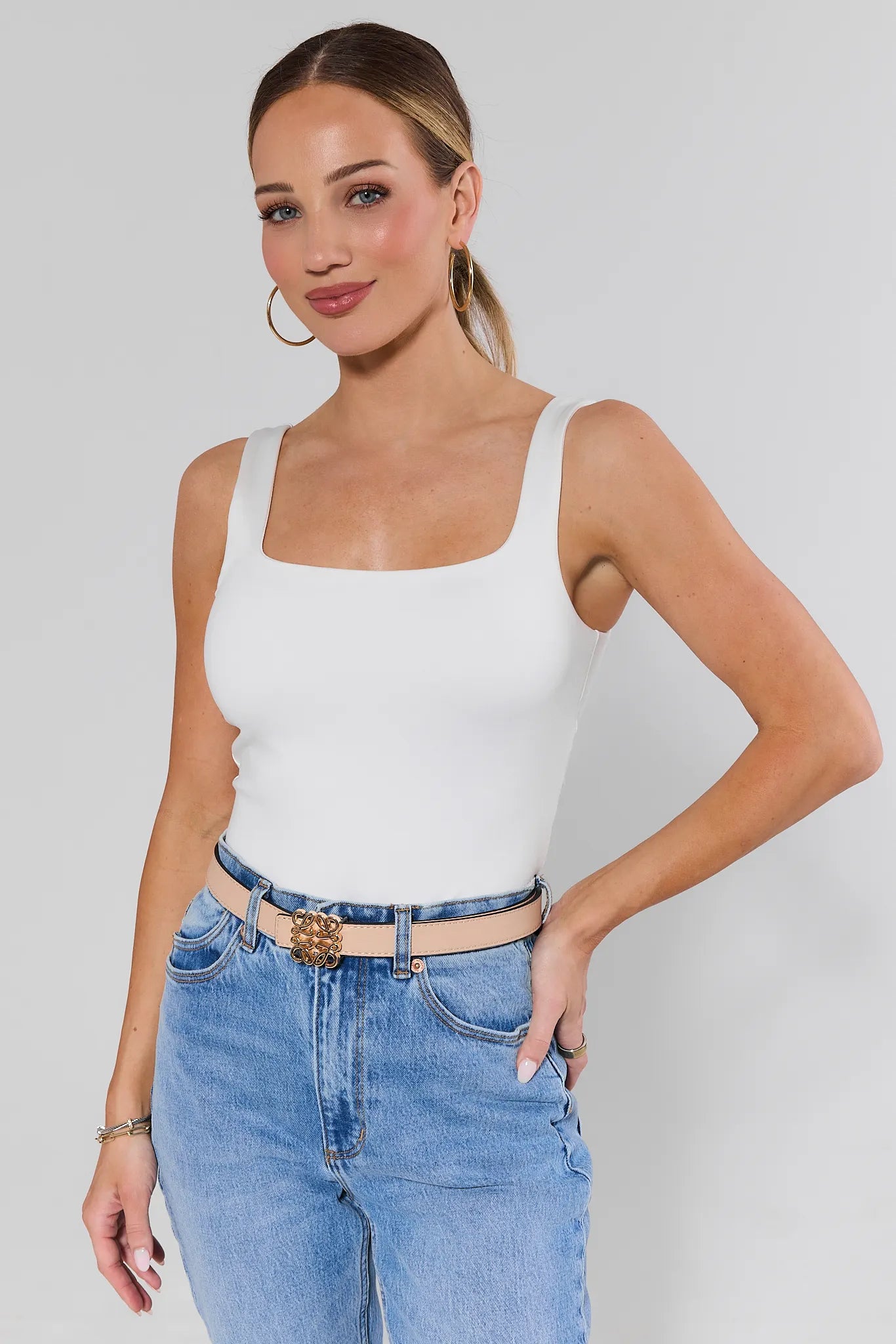 Woman wearing a white tank top and blue jeans and rowan belt against a plain background