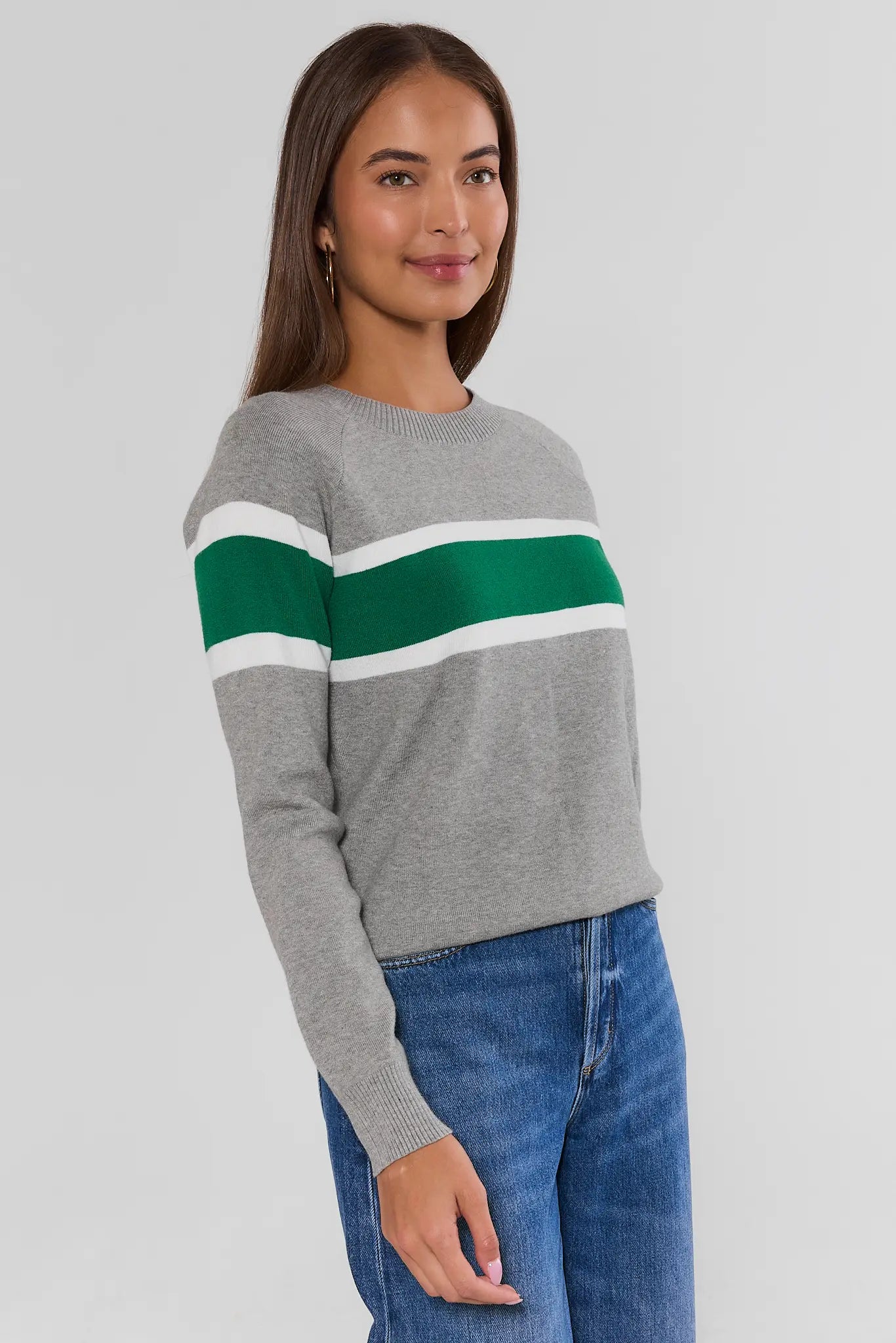 A woman wearing a gray crewneck sweater with a bold green and white horizontal stripe across the chest, paired with blue jeans, photographed against a light gray studio background.