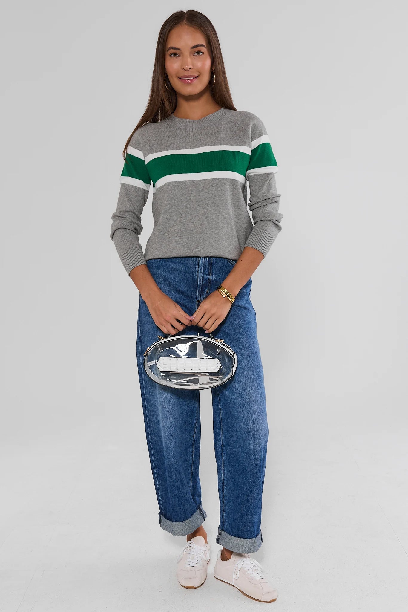 A woman modeling a gray crewneck sweater with green and white horizontal stripes, paired with blue jeans and white sneakers, holding a clear clutch with a white airplane design against a light gray studio background.