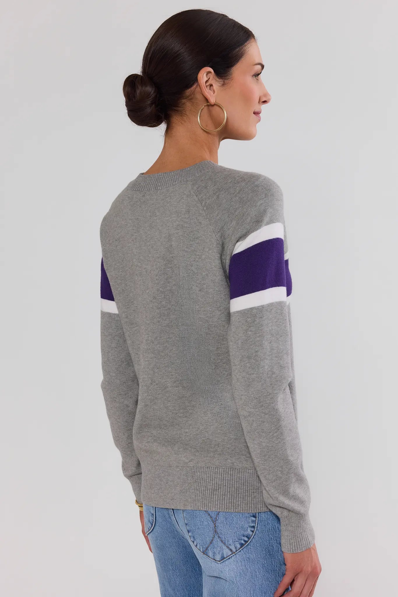 A woman shown from the back wearing a heather gray knit sweater with purple and white stripe details on the upper arms, paired with blue jeans, against a plain light gray studio background.