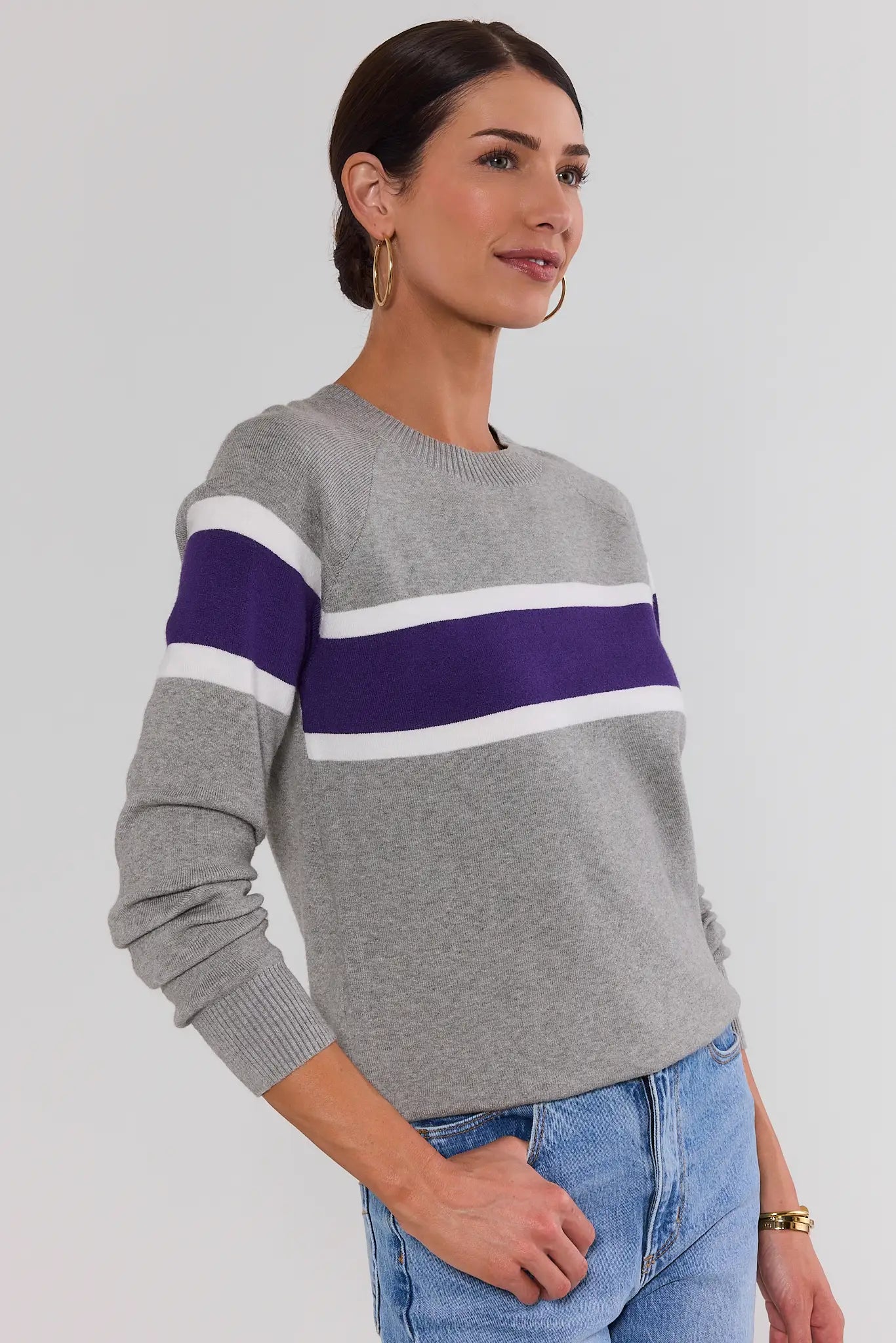 A woman wearing a gray knit sweater with a wide purple and white horizontal stripe across the chest, paired with blue jeans, photographed against a light gray studio background.