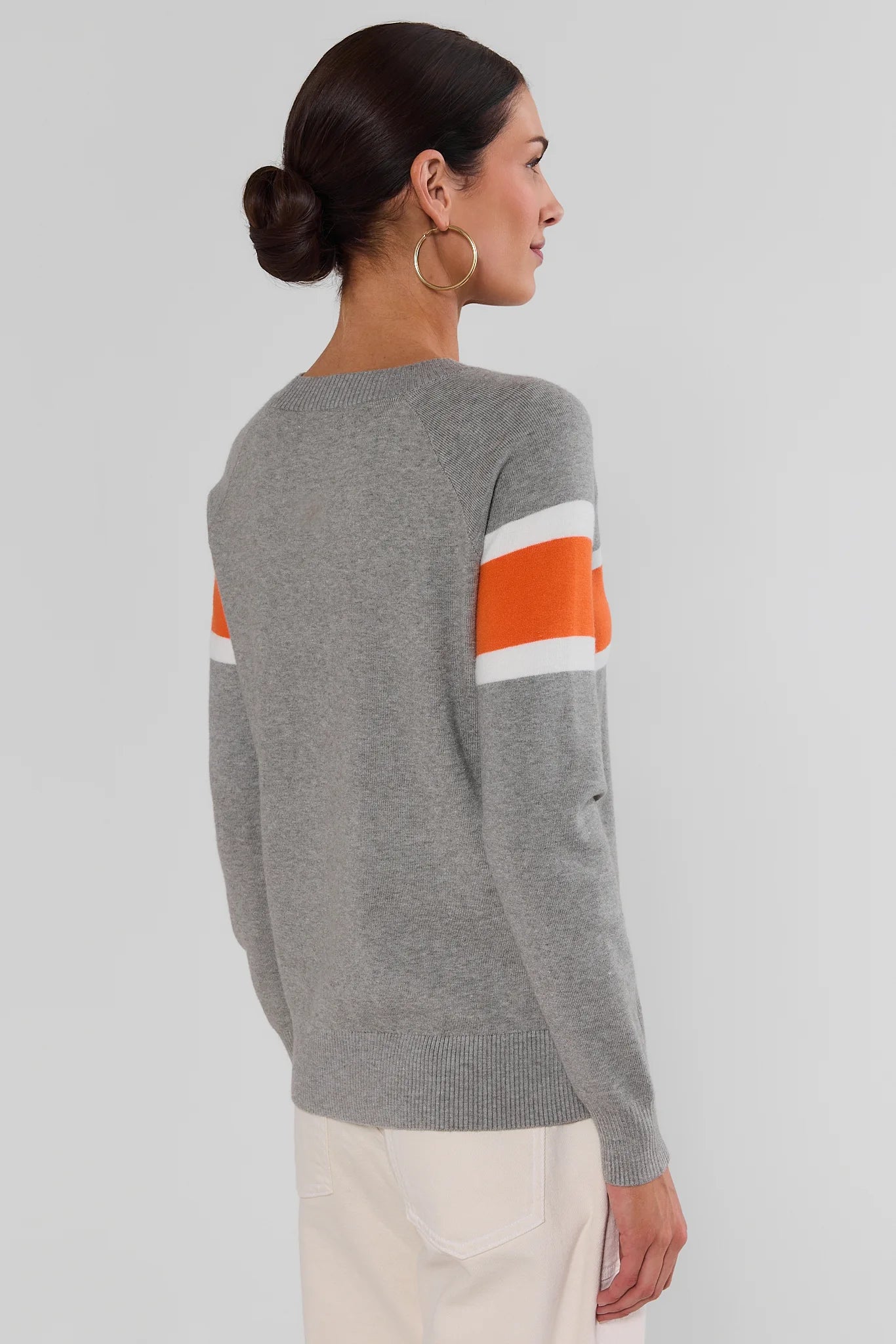 A woman shown from the back wearing a heather gray knit sweater with orange and white stripes on the upper sleeves, paired with cream pants, against a light gray studio background.