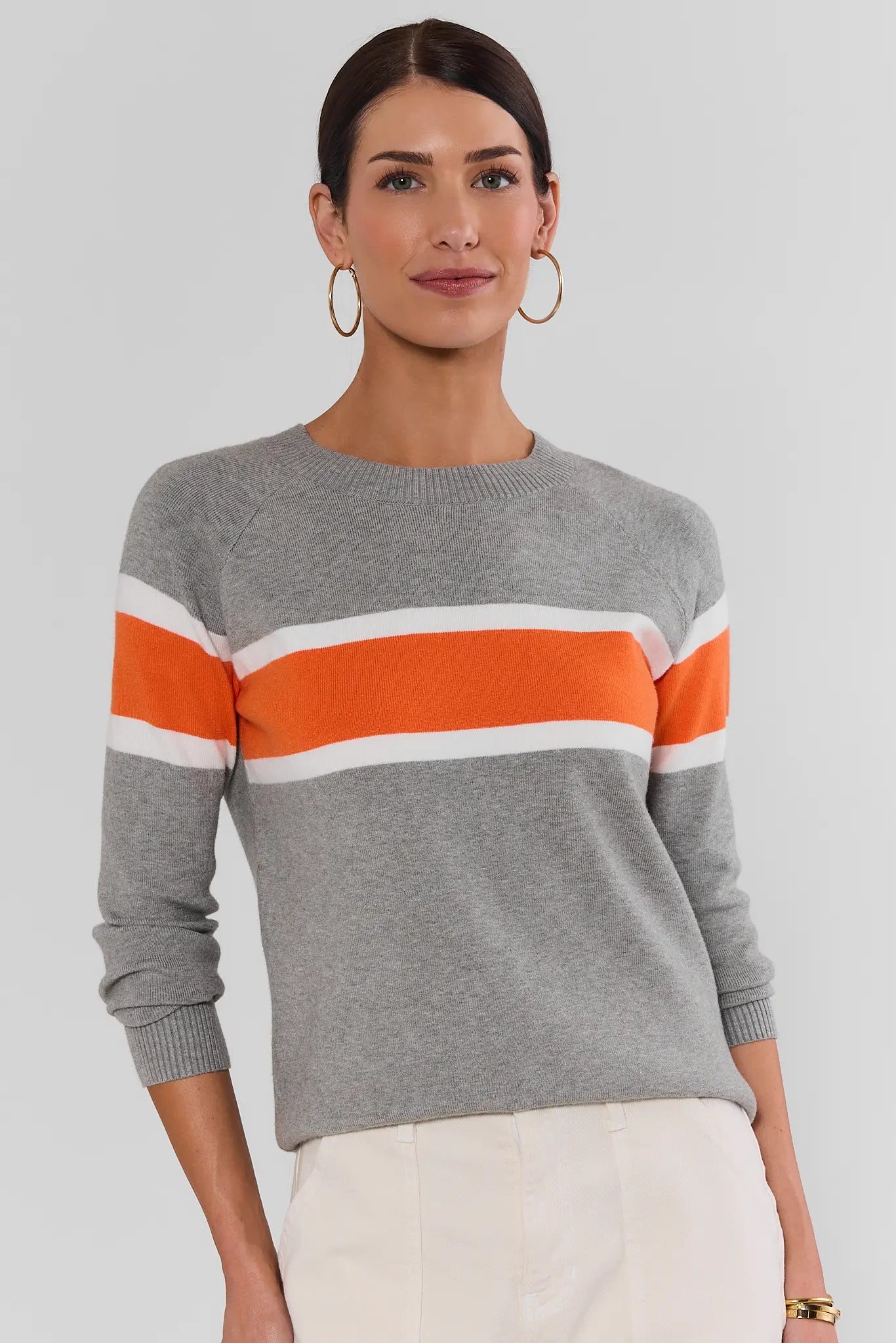 A woman wearing a gray knit sweater with bold horizontal orange and white stripes across the chest and shoulders, paired with off-white pants, against a plain light gray studio background.