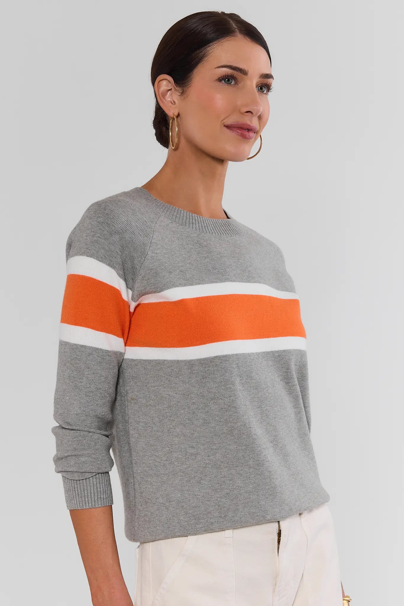 A woman wearing a heather gray knit sweater with a bold orange and white horizontal stripe pattern across the chest and upper arms, paired with off-white trousers, set against a plain light gray studio background.