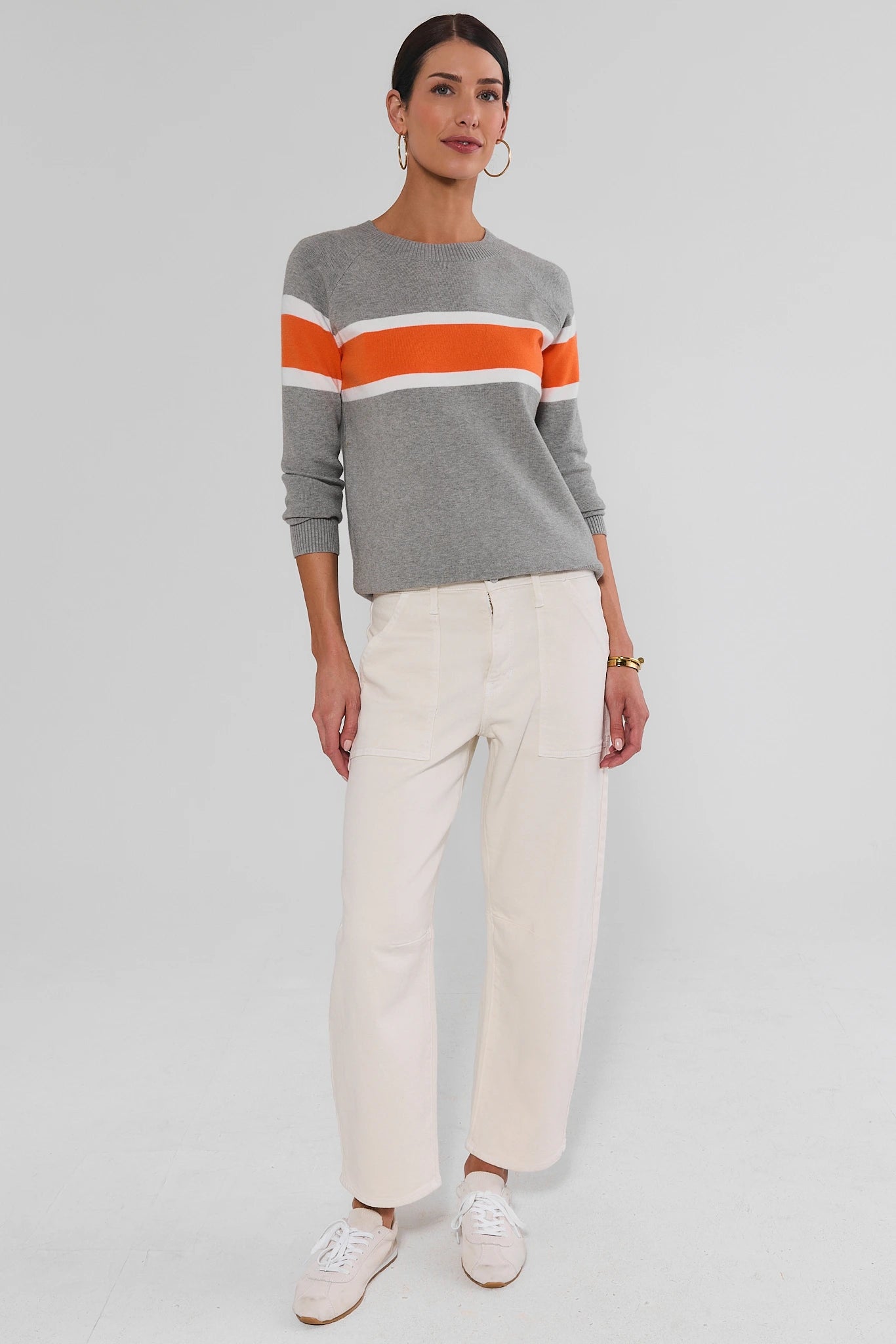 A woman modeling a gray crewneck sweater with horizontal orange and white stripes across the chest, paired with off-white trousers and white sneakers, against a light gray studio background.