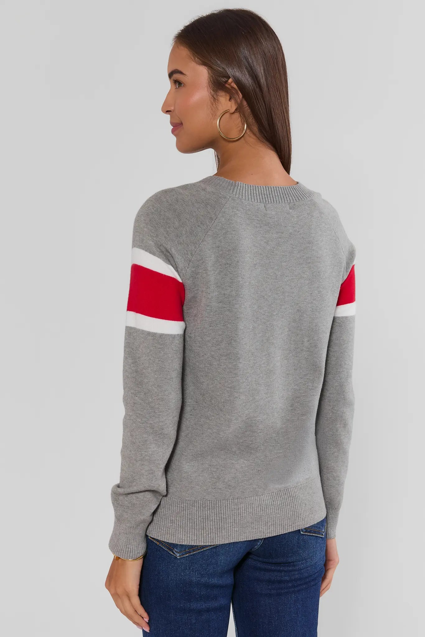A woman shown from the back wearing a heather gray knit sweater with red and white stripes on the upper sleeves, paired with blue jeans, against a light gray studio background.