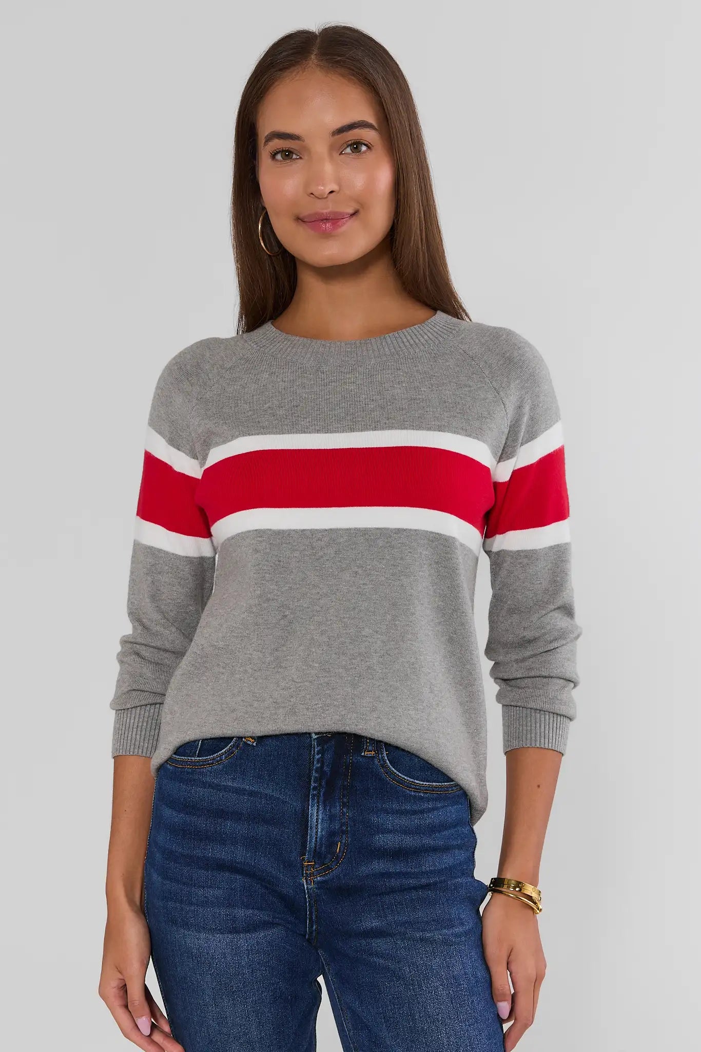 A woman wearing a gray knit sweater with bold red and white horizontal stripes across the chest and shoulders, paired with blue jeans, photographed against a light gray studio background.