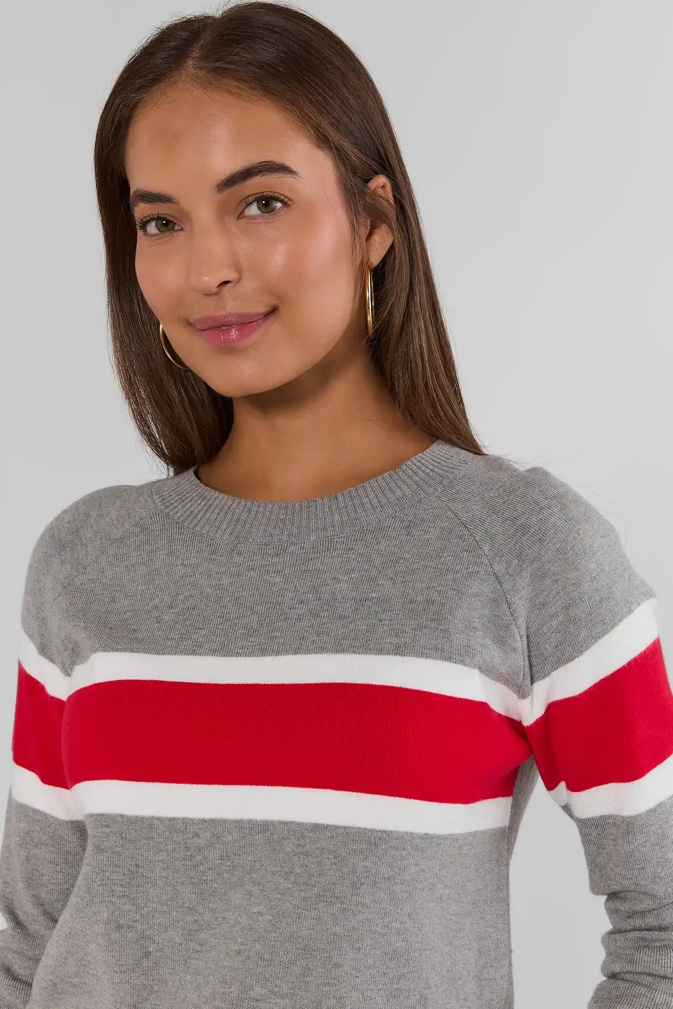 A woman wearing a gray crewneck sweater with bold horizontal red and white stripes across the chest and sleeves, photographed against a light gray studio background.