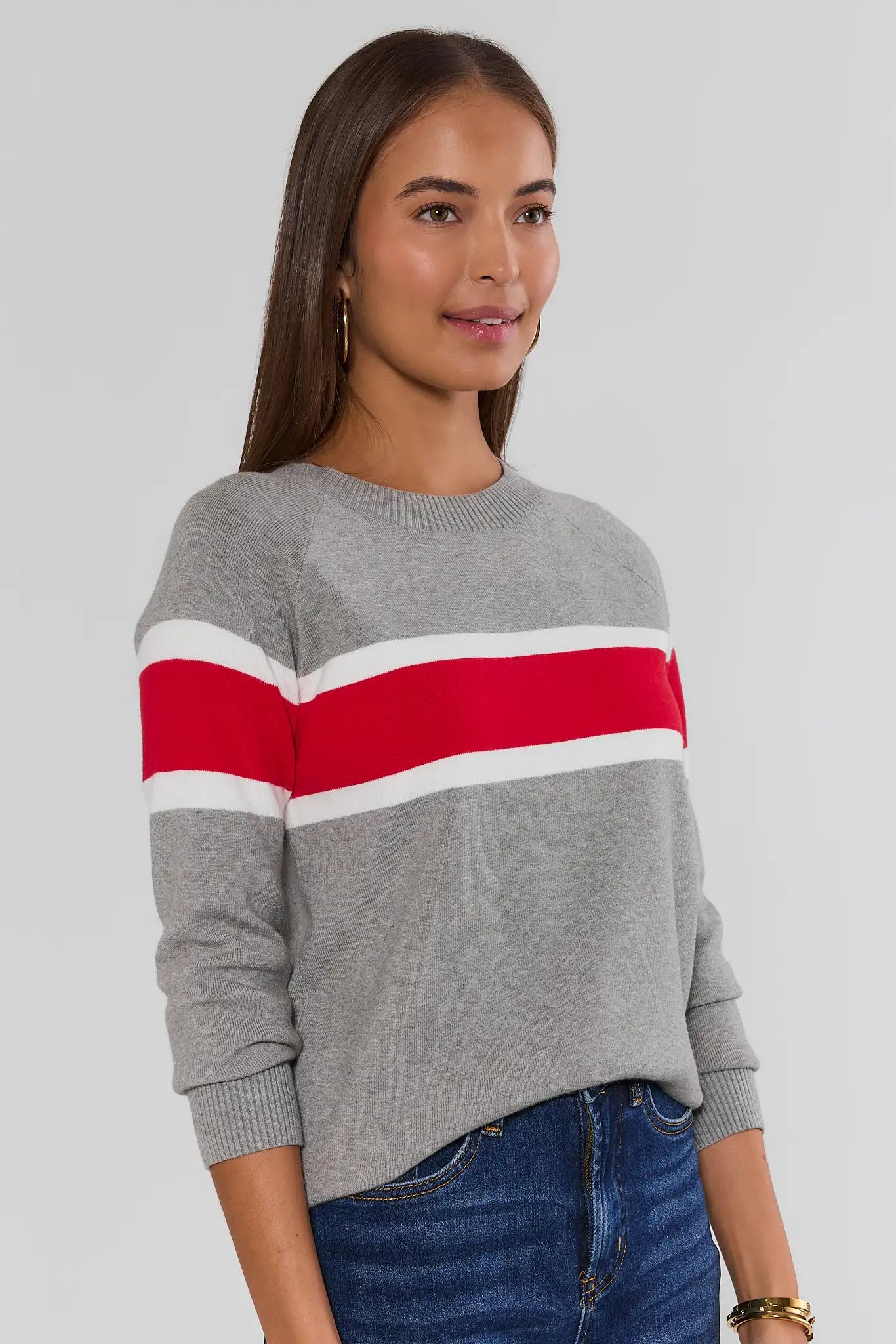 A woman wearing a gray knit sweater with a bold horizontal stripe pattern in red and white, paired with blue jeans, against a solid light gray background.