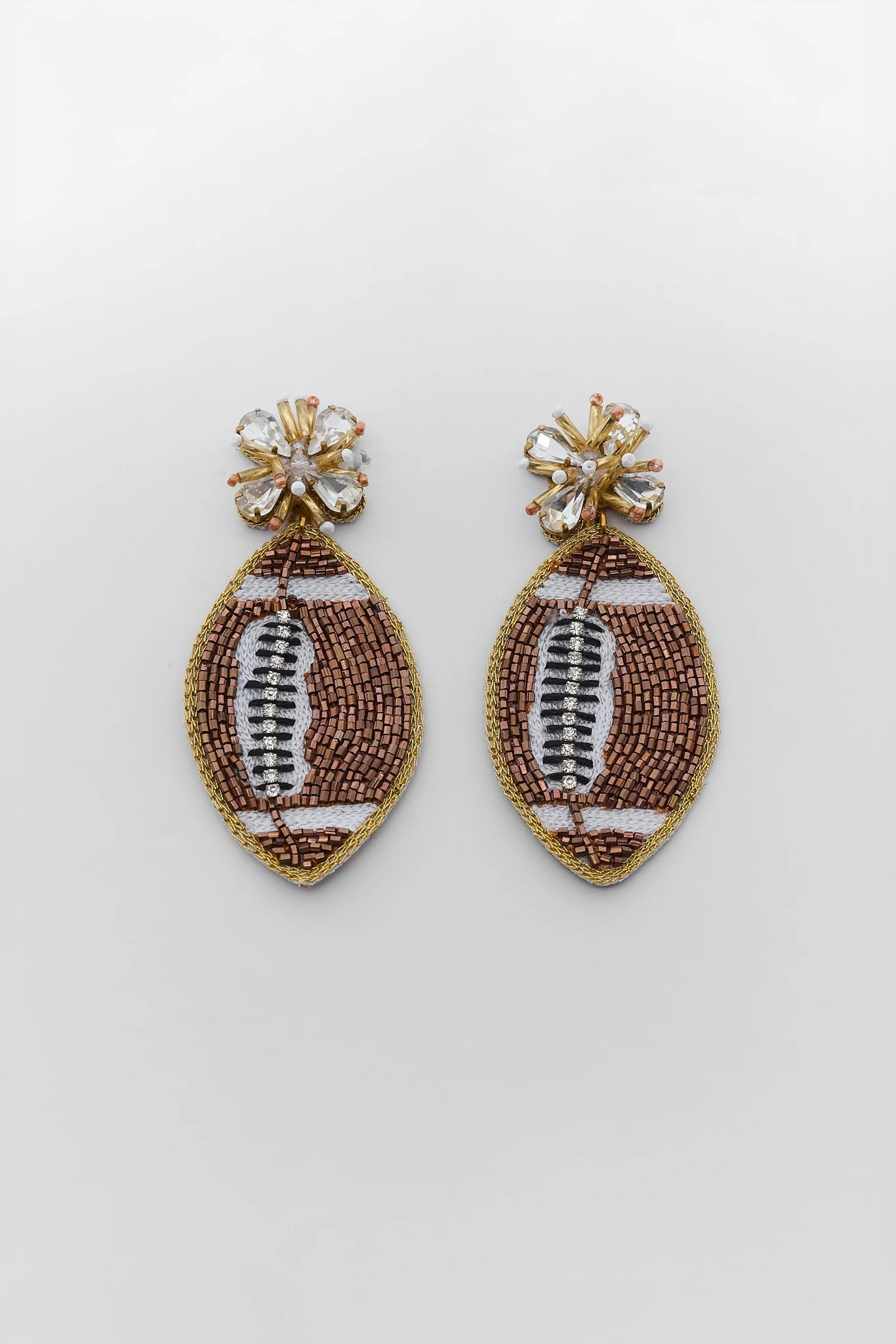 A pair of gold-toned beaded earrings shaped like footballs, featuring brown and white beadwork with clear rhinestone studs at the top, shown on a light gray background.
