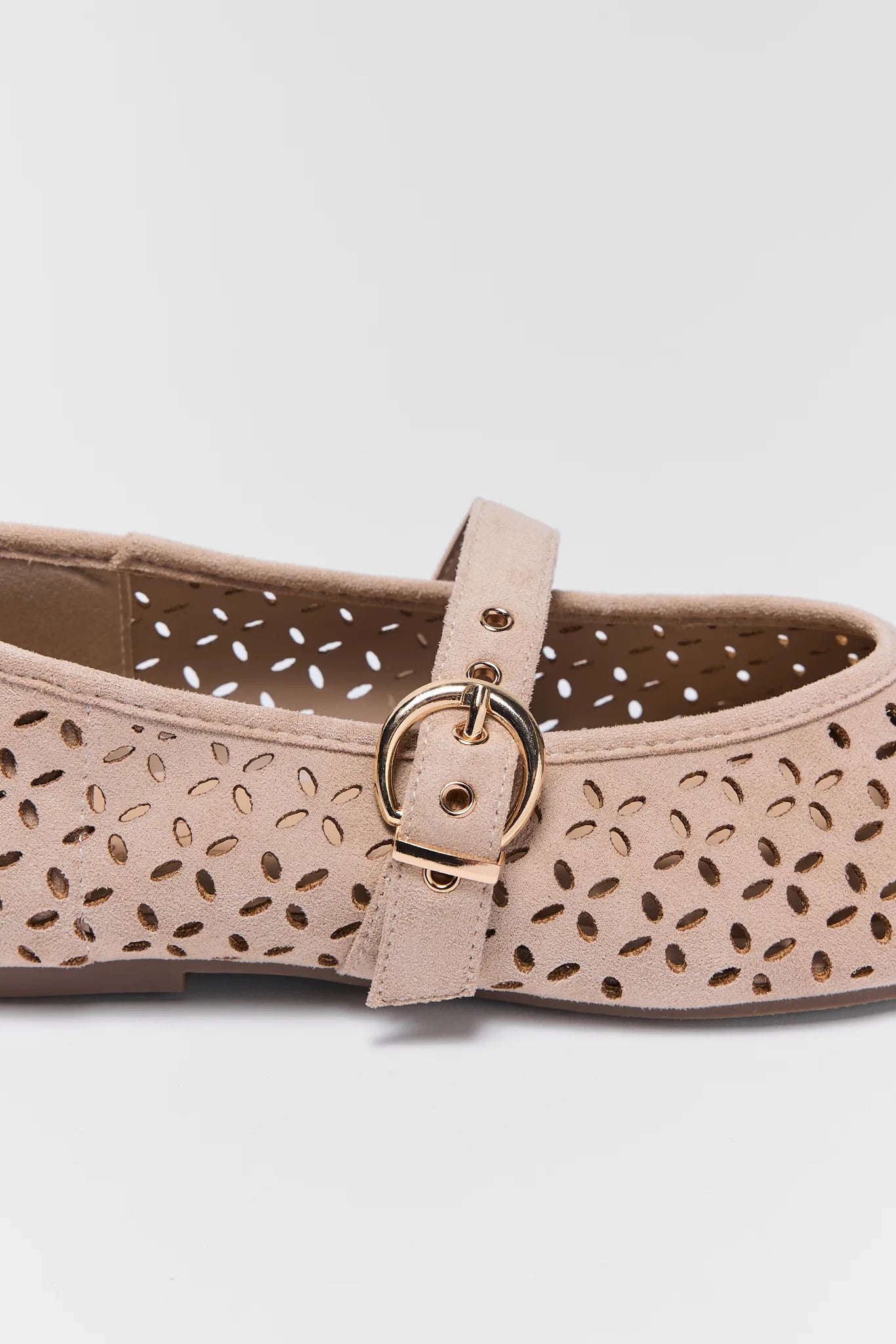 A close-up of a beige flat shoe with an allover leaf-shaped laser cutout pattern and a matching beige strap featuring a gold-tone metal buckle.